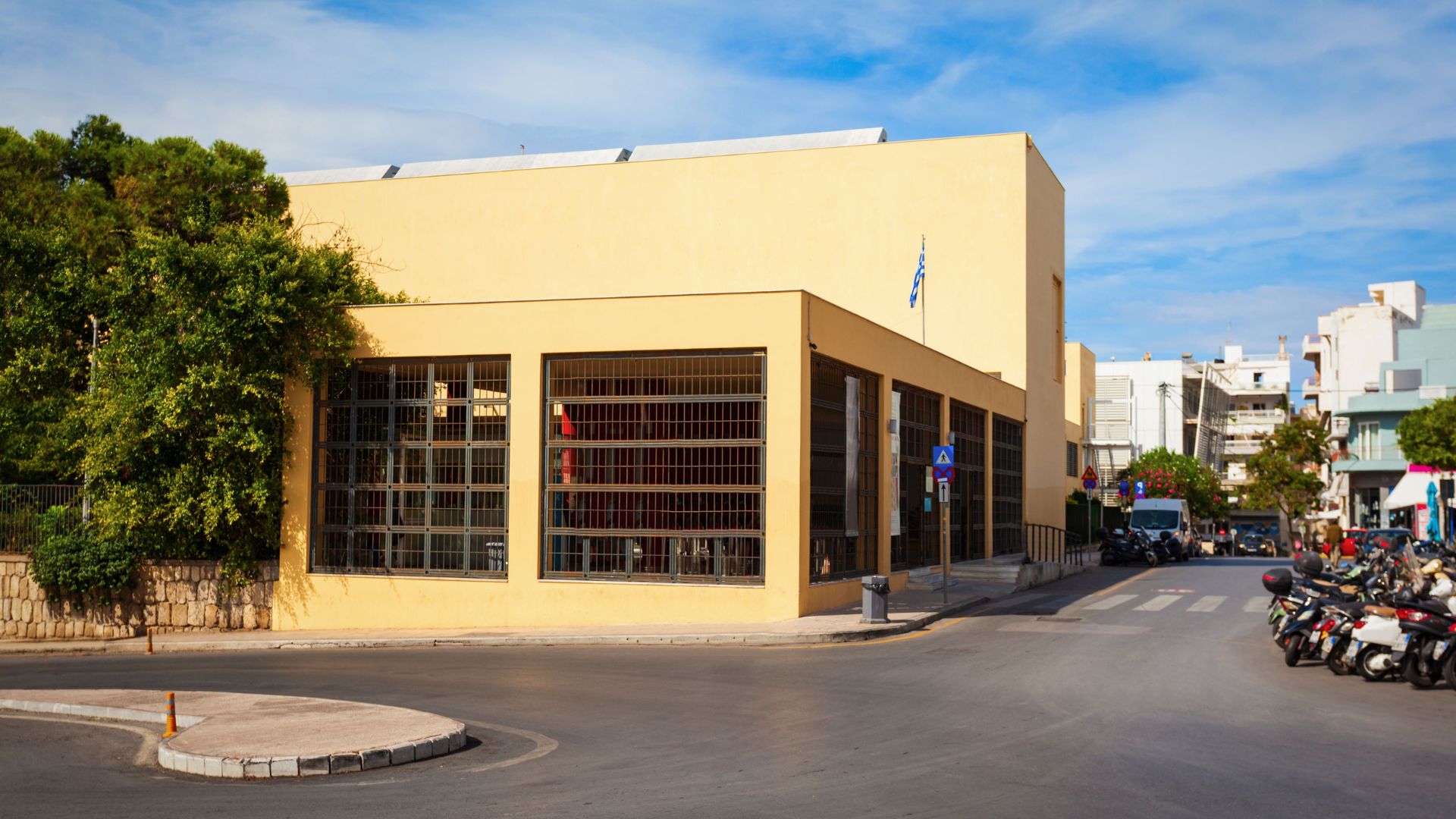 Exterior view of the Heraklion Archaeological Museum, a yellow building with large windows, situated on a street corner in Heraklion, Crete, with scooters parked nearby.