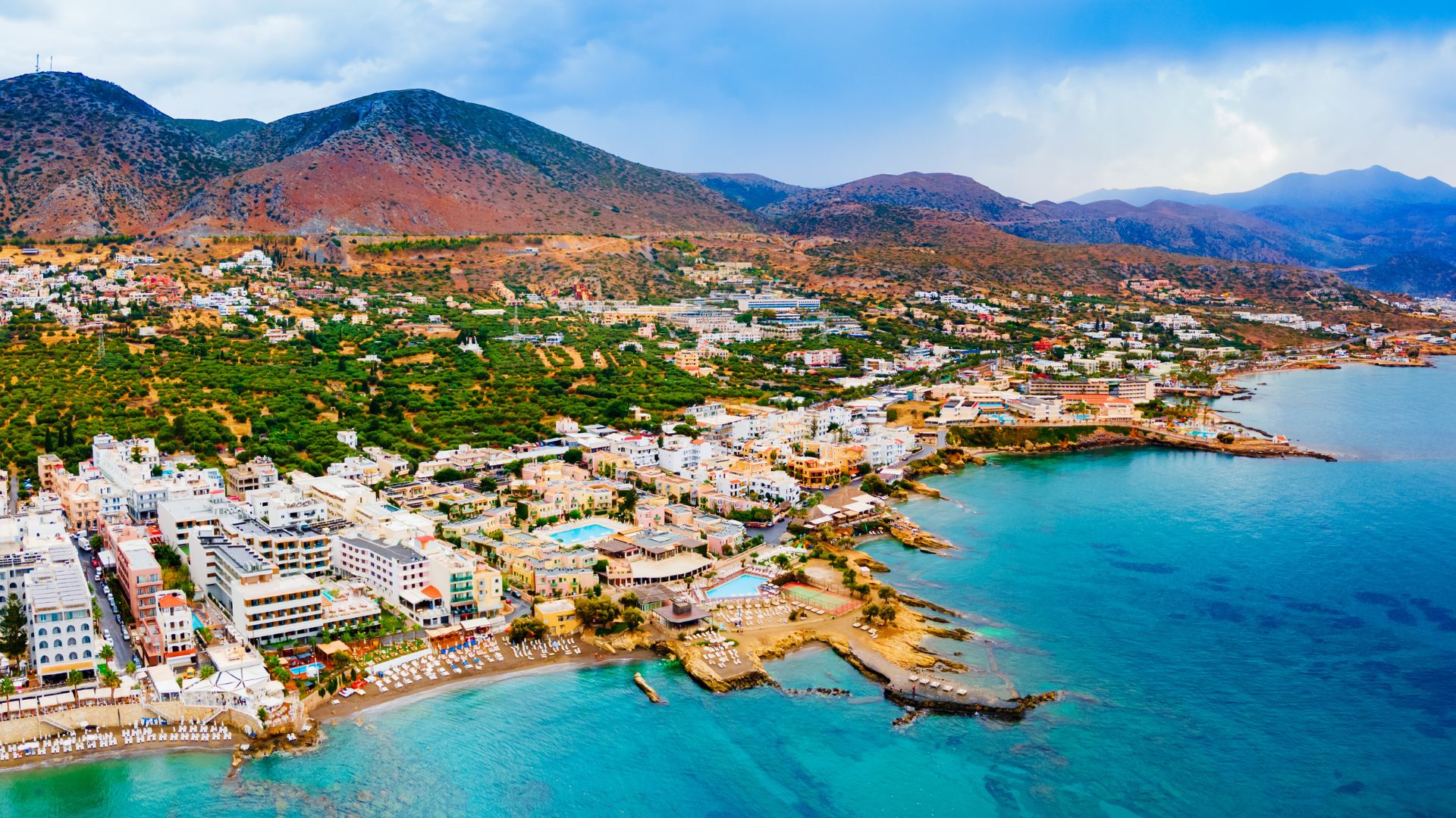 Aerial view of Hersonissos, Crete, featuring the coastline, town buildings, and surrounding mountains.