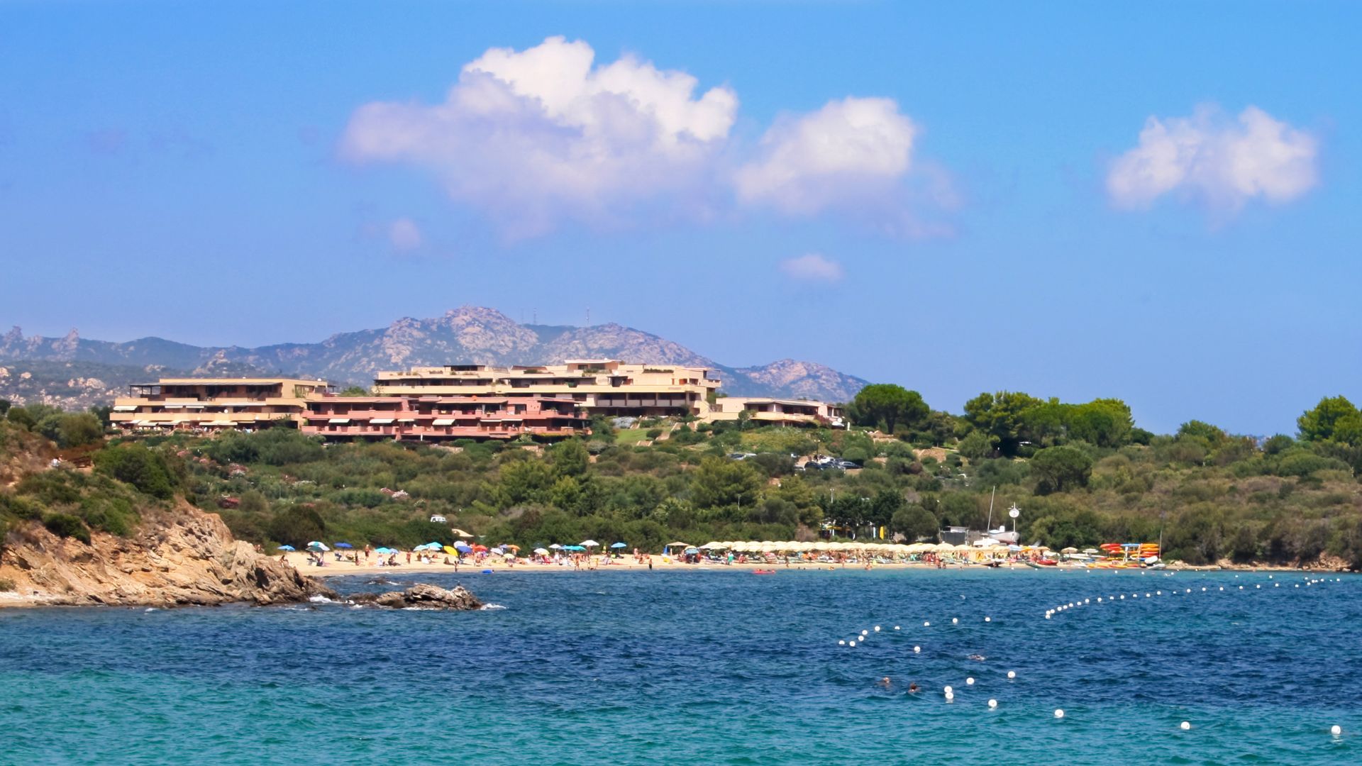 The Hotel Cala di Volpe in Porto Cervo, Sardinia, Italy, is nestled on a picturesque bay with clear turquoise water and a sandy beach, surrounded by lush greenery and rocky hills