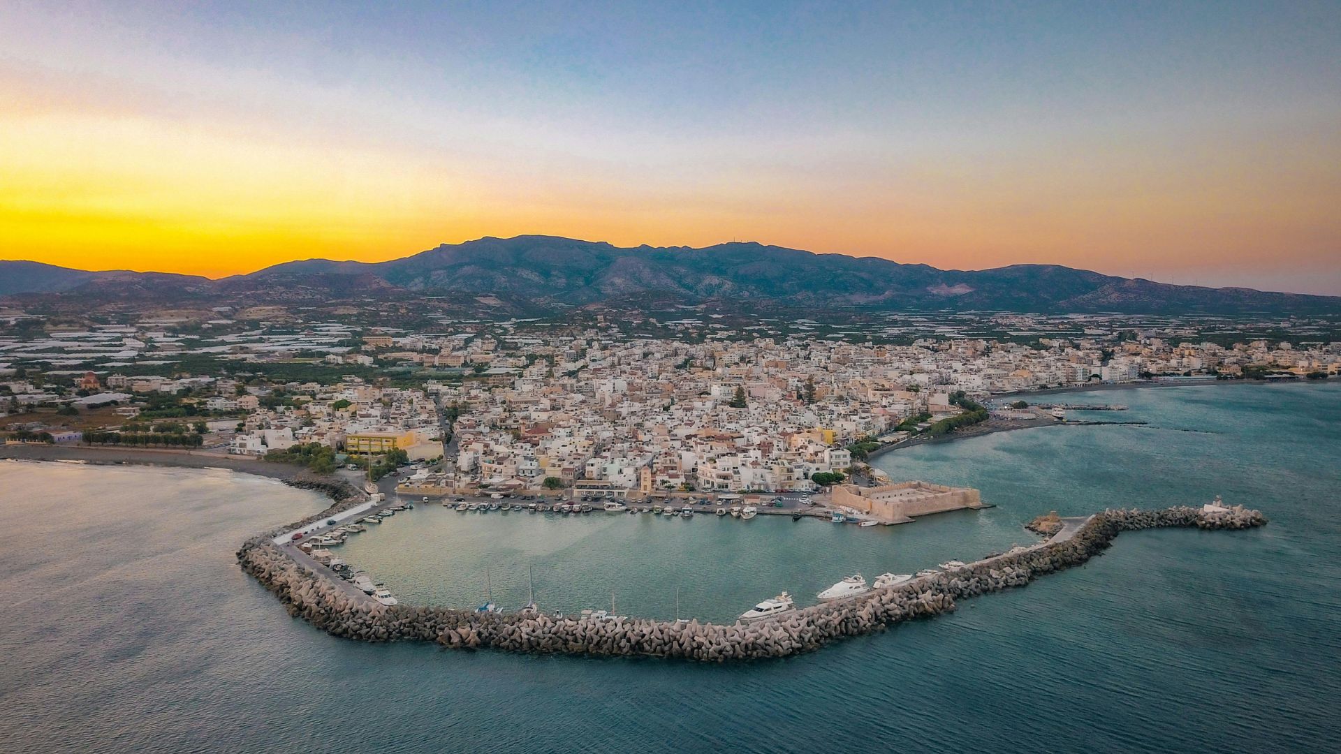 Aerial view of Ierapetra, Crete, with a harbor and city buildings at sunset.