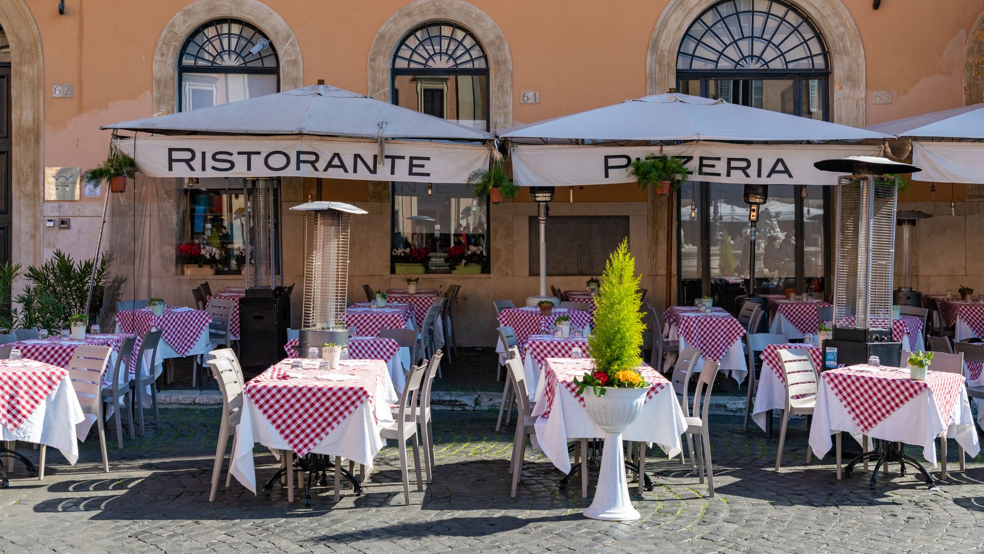 Outdoor seating area of 'Ristorante Pizzeria' in Rome, featuring tables with red and white checkered cloths, outdoor heaters, and a lemon cypress centerpiece.