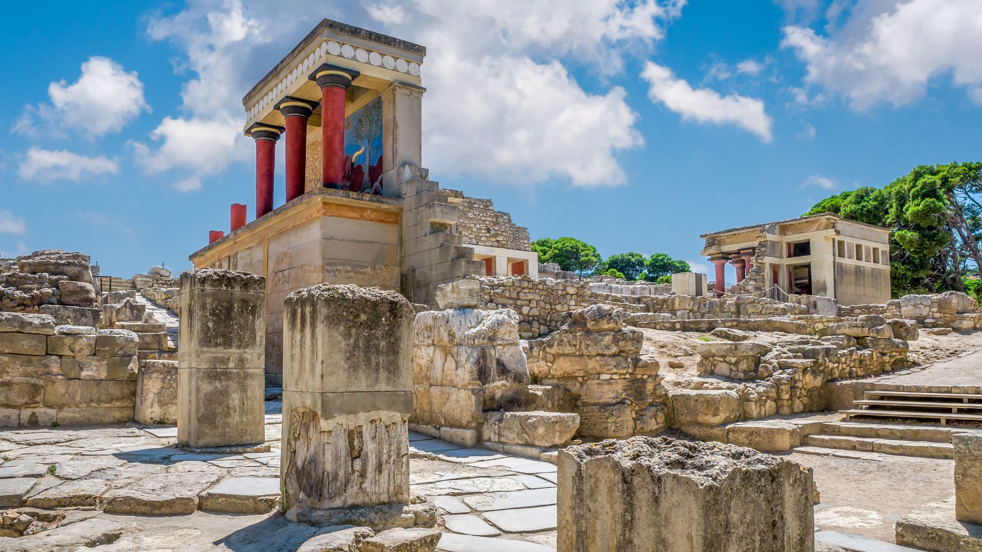 Ruins of the ancient Minoan palace of Knossos in Crete, featuring stone structures and columns under a blue sky.