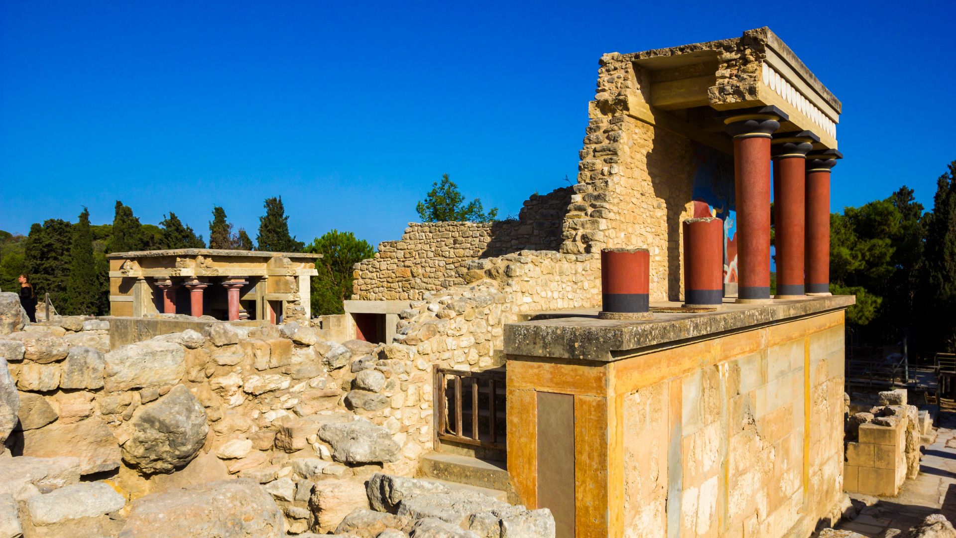 Ruins of the ancient Minoan palace of Knossos in Crete, featuring stone structures and columns under a blue sky.