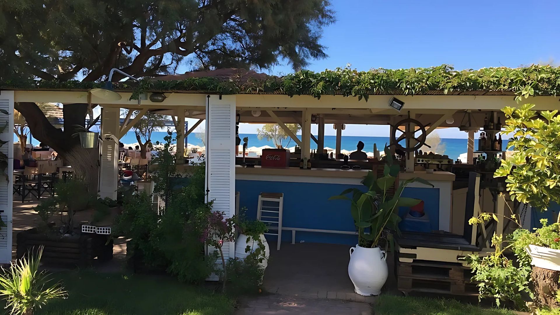 The image shows the open-air Kyma Restaurant in Plakia, Greece, situated along the beach. The restaurant features a wooden structure with a roof covered in greenery, providing shade for the dining area and bar.