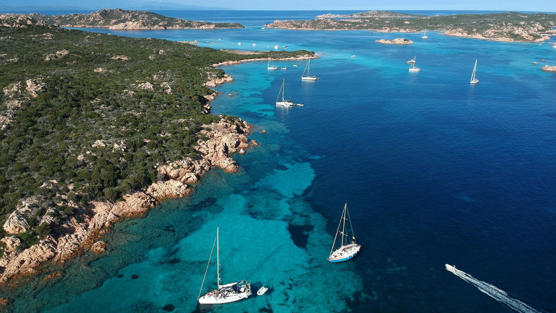 Aerial view of blue waters, rocky shore, greenery, and some sailboats at La Maddalena in Sardinia, Italy.