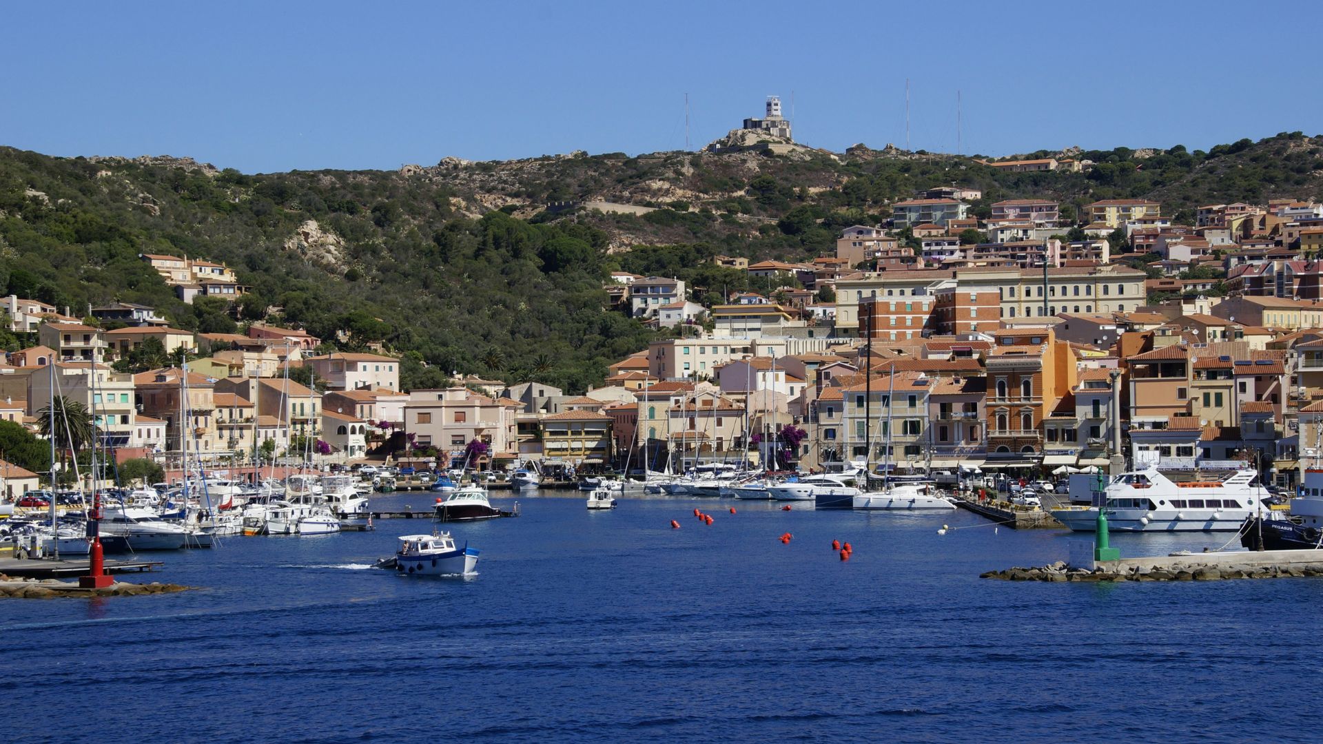 Town view of La Maddalena in Sardinia characterized by buildings, mountains, greenery, blue waters, and boats.