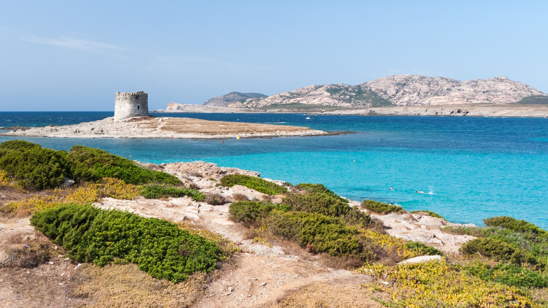 A scenic view of La Pelosa Beach in Sardinia, featuring turquoise water, white sand, and a historic tower
