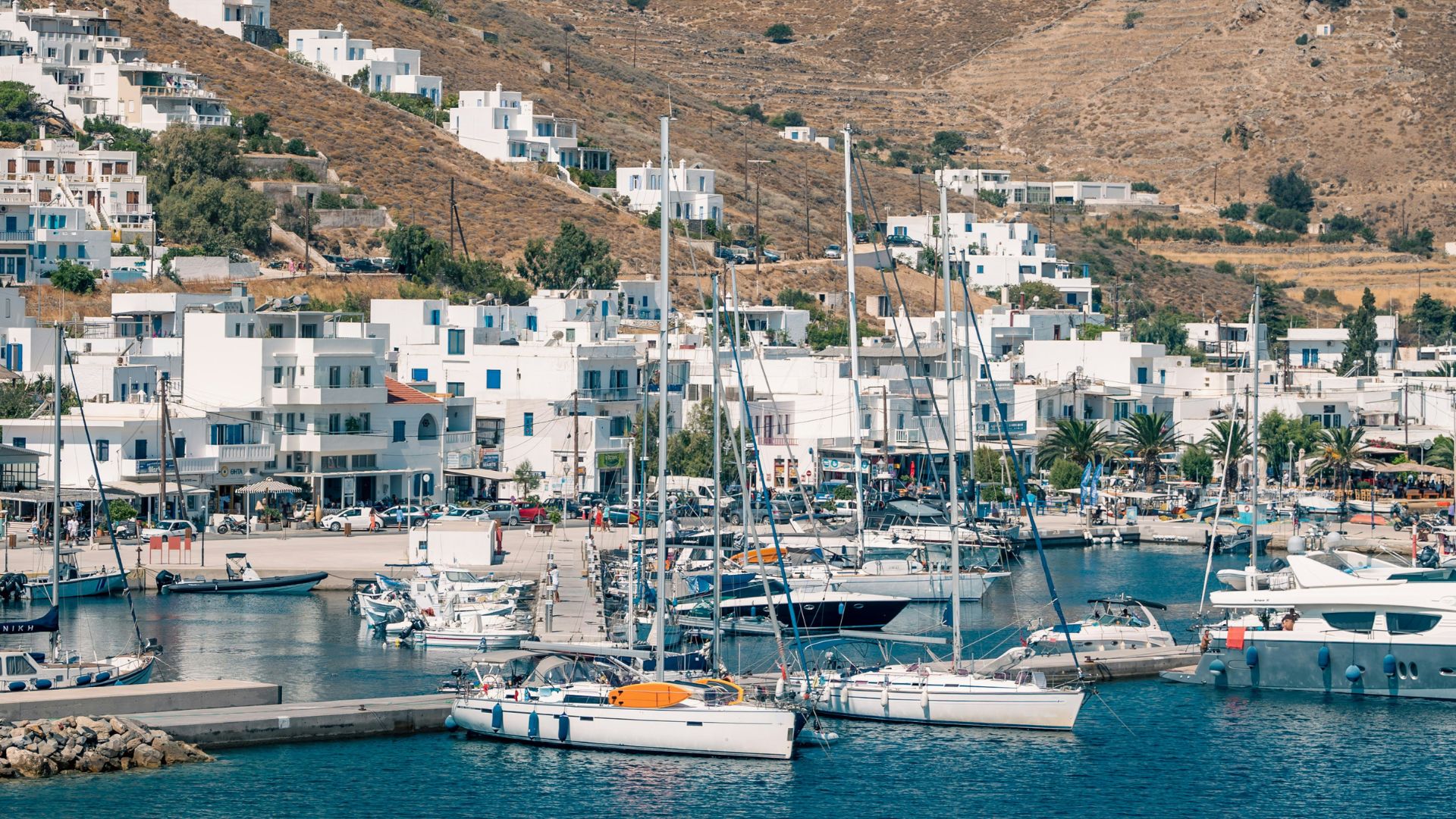 View of the port of Livadi in Serifos, Greece, with white buildings and docked boats against a mountainous backdrop.