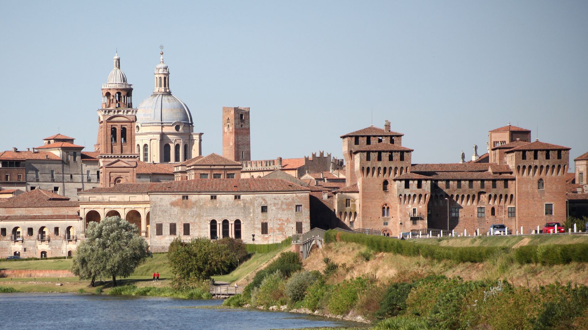 The Palazzo Ducale in Mantua, Italy, a large complex of buildings with towers and courtyards, seen from across a river.