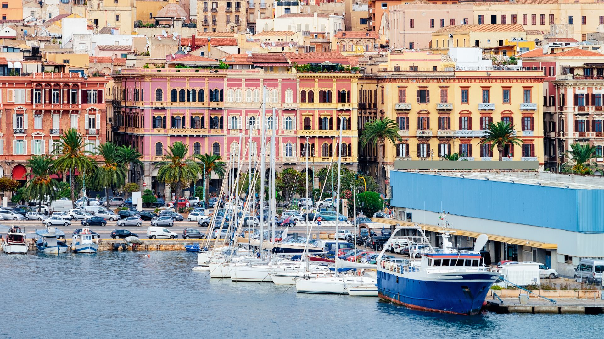 A view of the Marina di Cagliari in Sardinia, Italy, featuring docked boats and colorful buildings in the background.