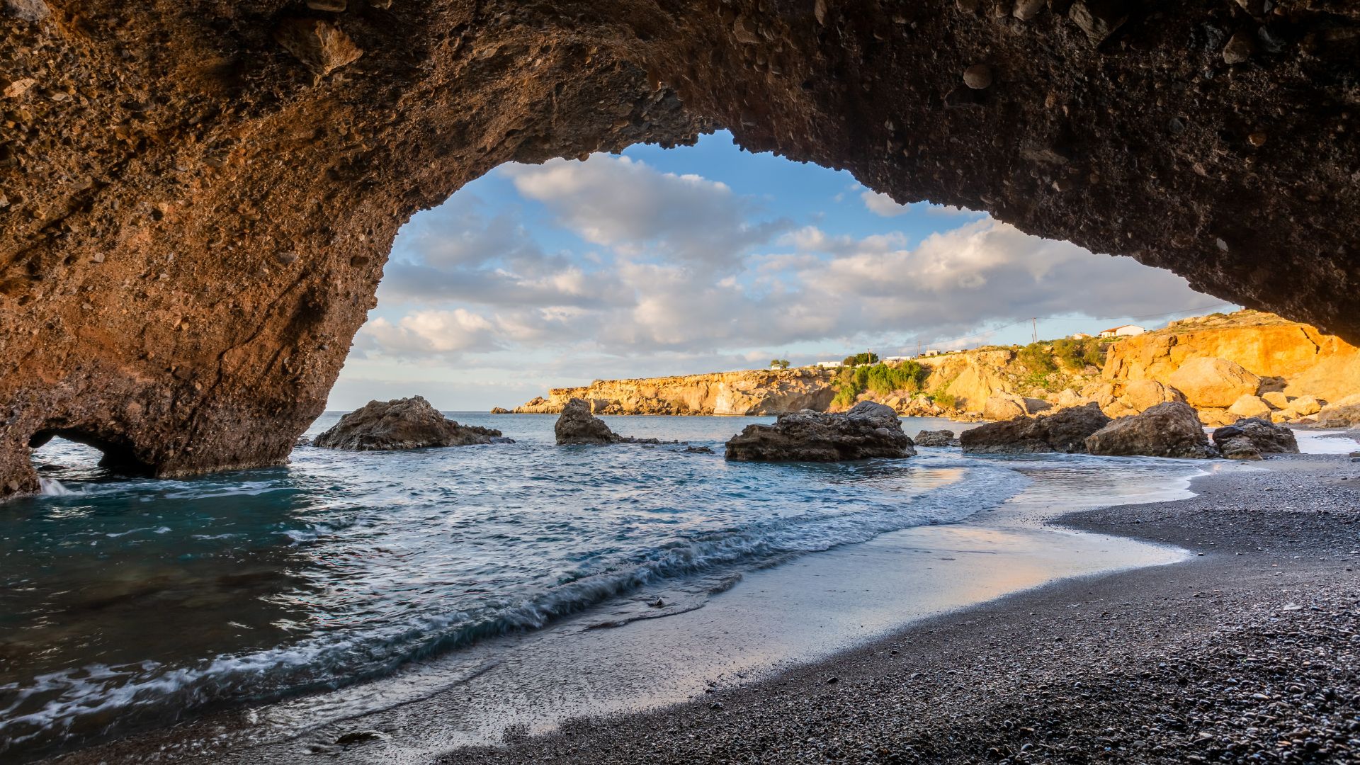 A view from inside a sea cave looking out at Matala Beach, with cliffs and rock formations in the distance under a partly cloudy sky.