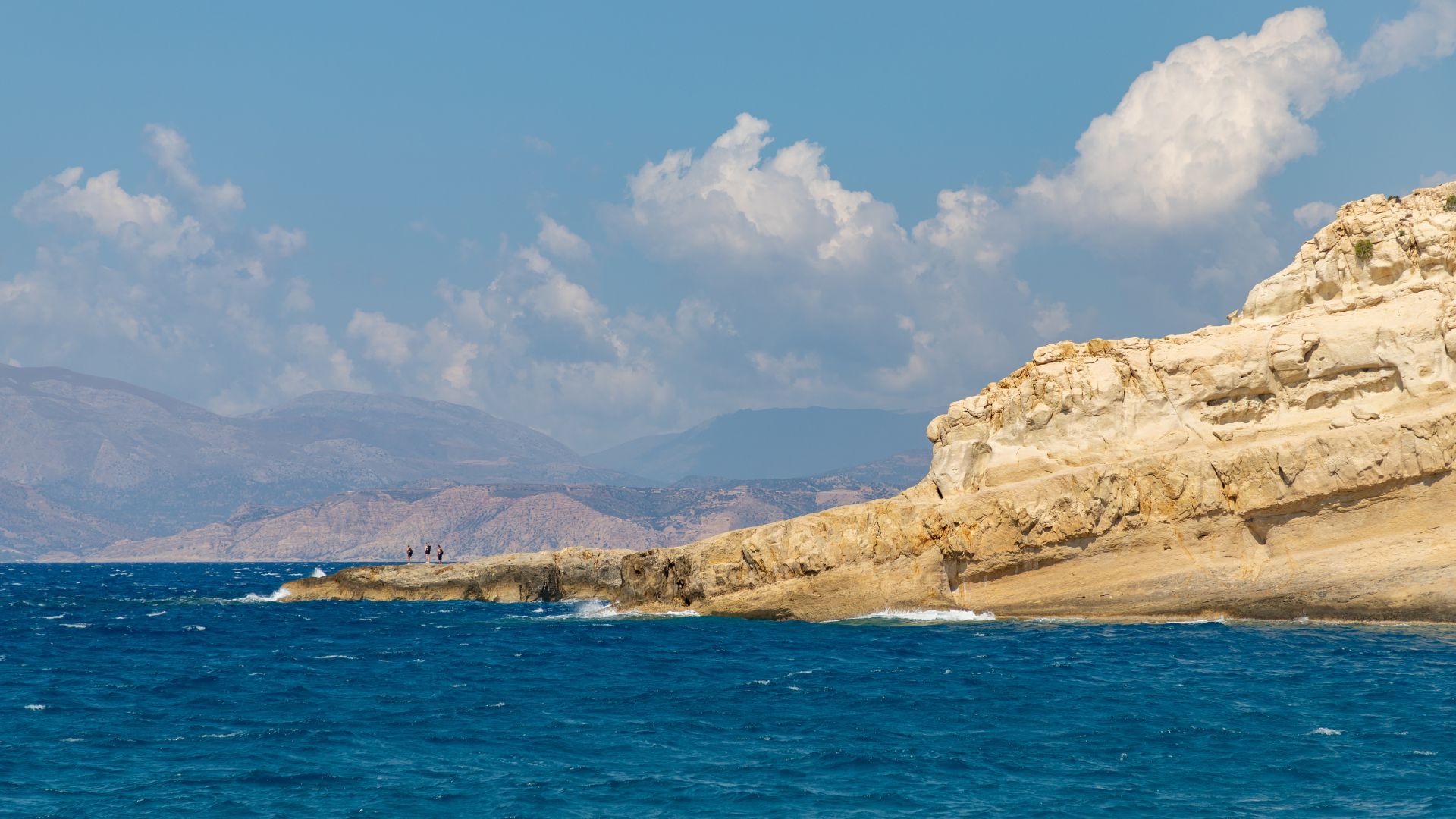 Matala Beach with its distinctive rock formations and caves, sandy shore, and clear blue waters under a partly cloudy sky.