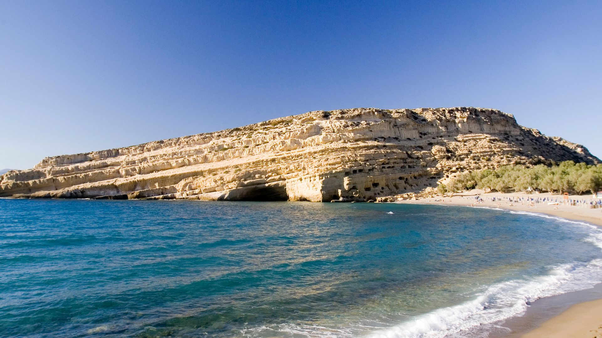 Matala Beach, Crete, featuring a sandy beach, blue water, and a cliff with caves.