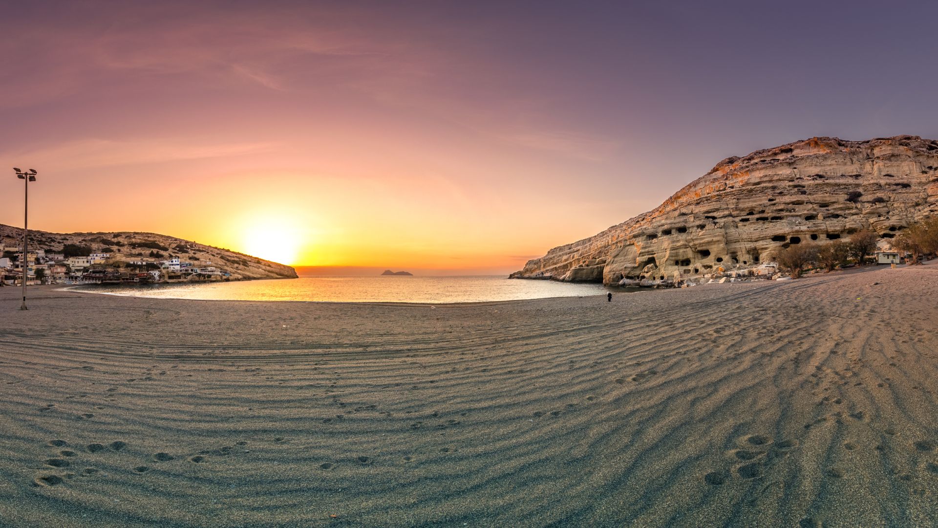 Panoramic view of Matala Beach at sunset, with caves in the cliffs and a sandy shoreline.