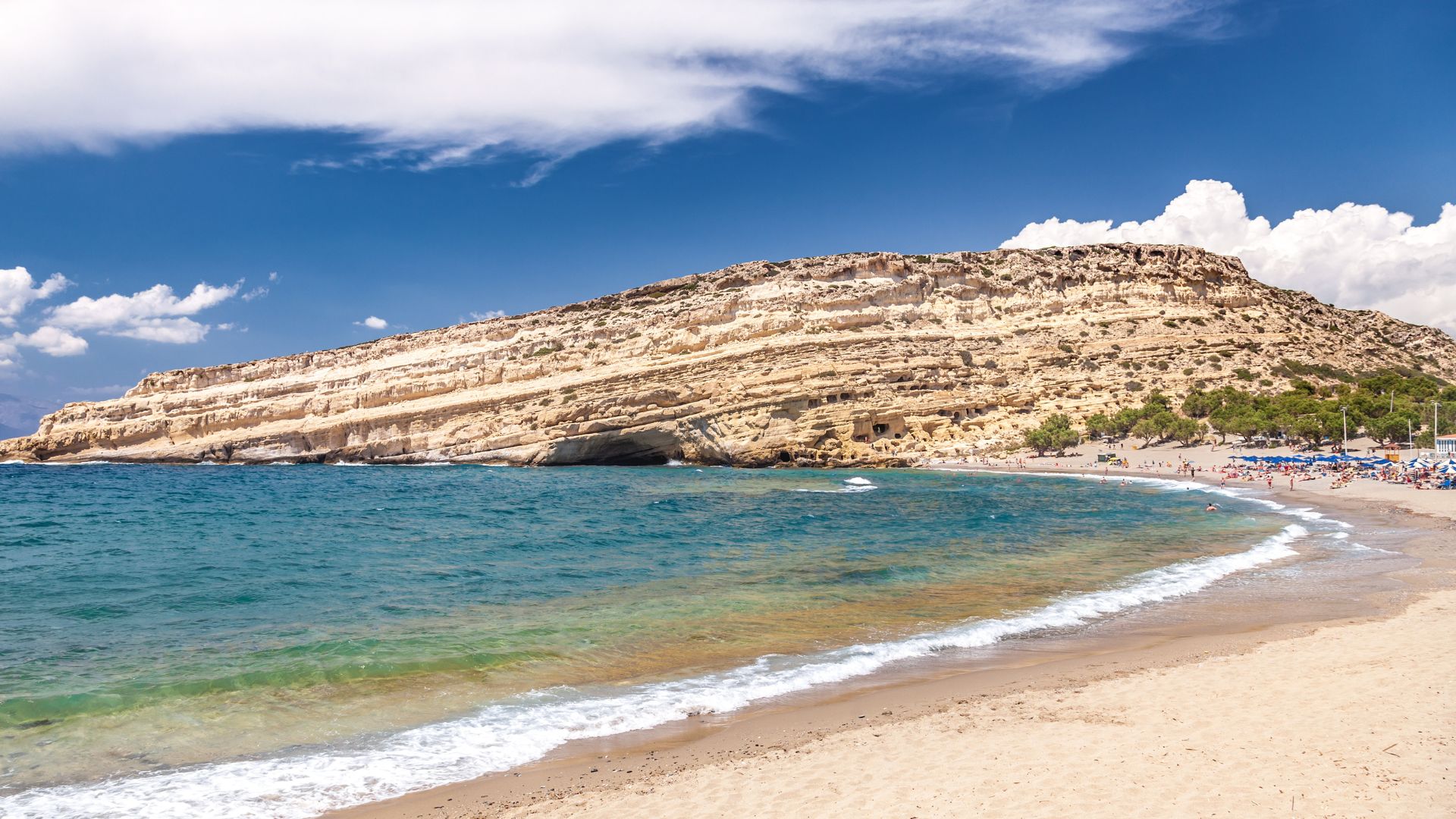 Matala Beach with its distinctive rock formations and caves, sandy shore, and clear blue waters under a partly cloudy sky.