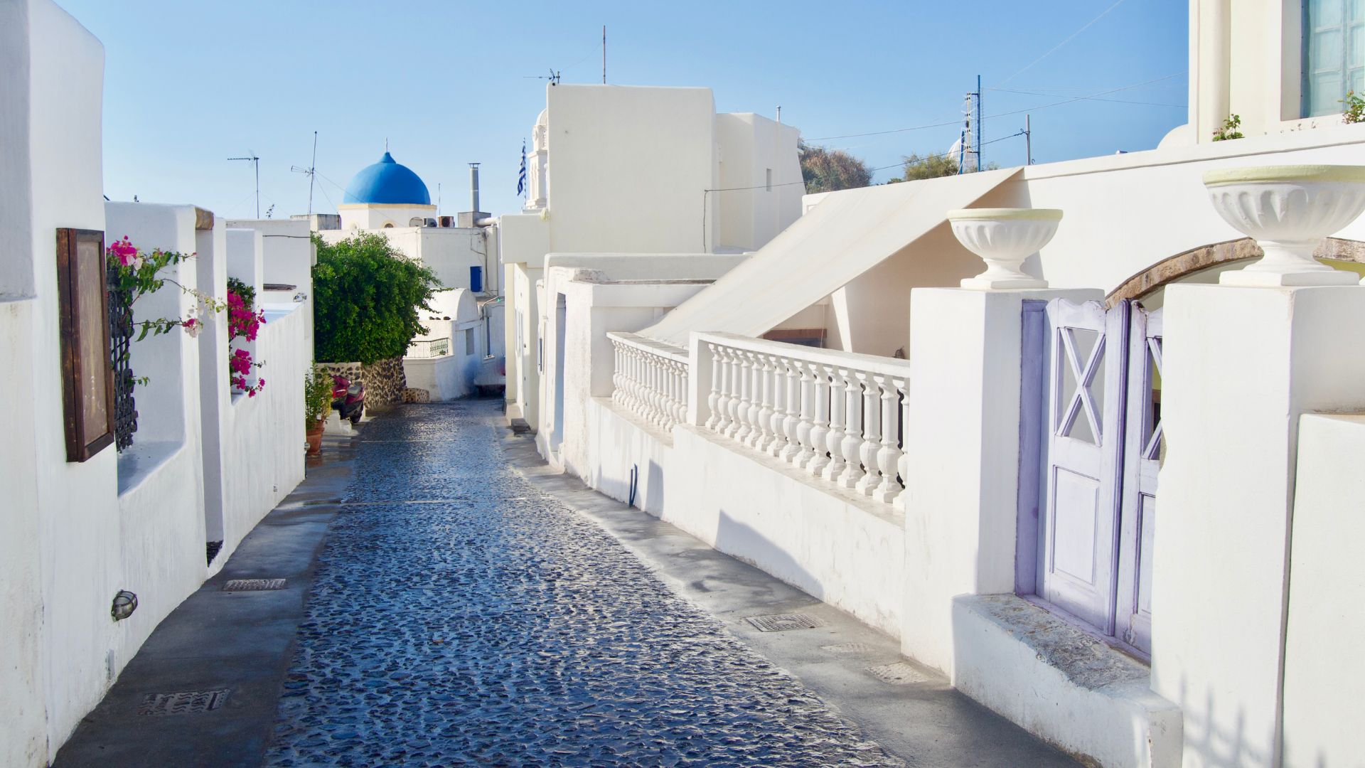 A narrow cobblestone street in Megalochori, Santorini, with white buildings and a blue-domed church in the background.