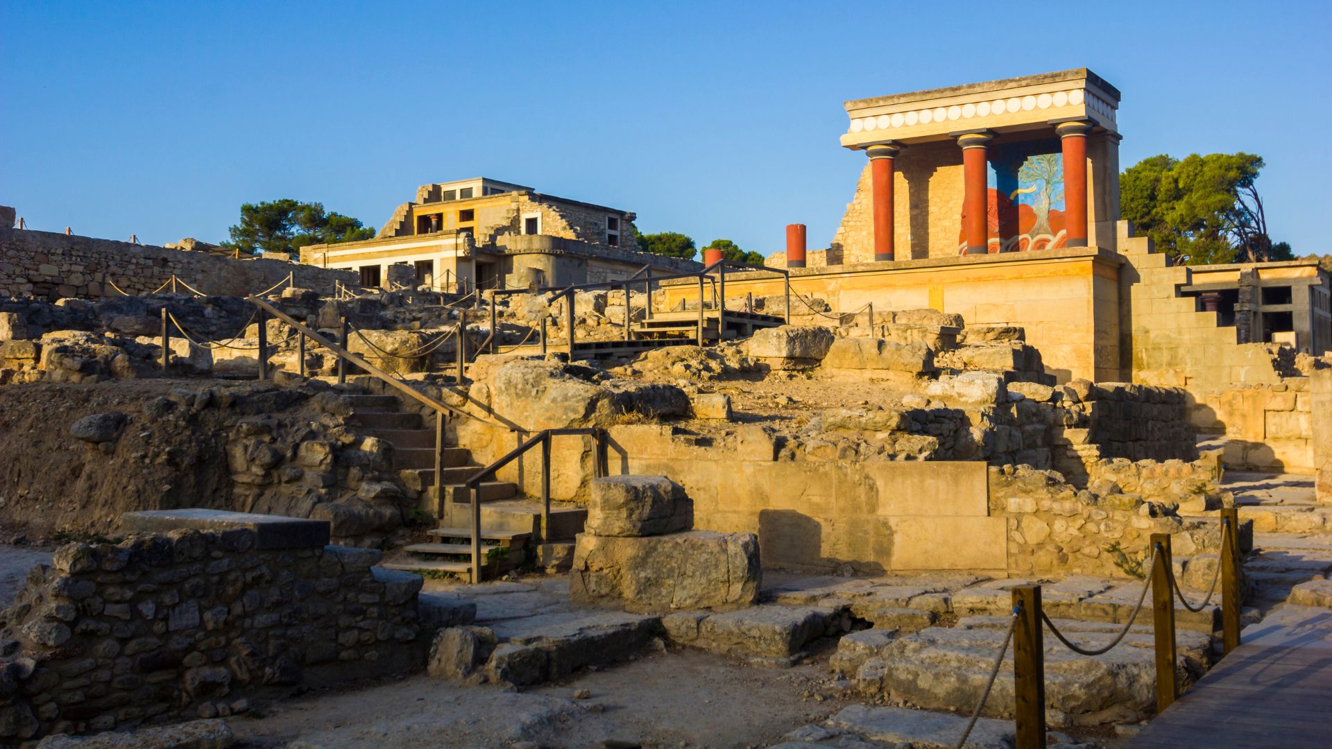 Ruins of the ancient Minoan palace of Knossos in Crete, featuring stone structures and columns under a blue sky.