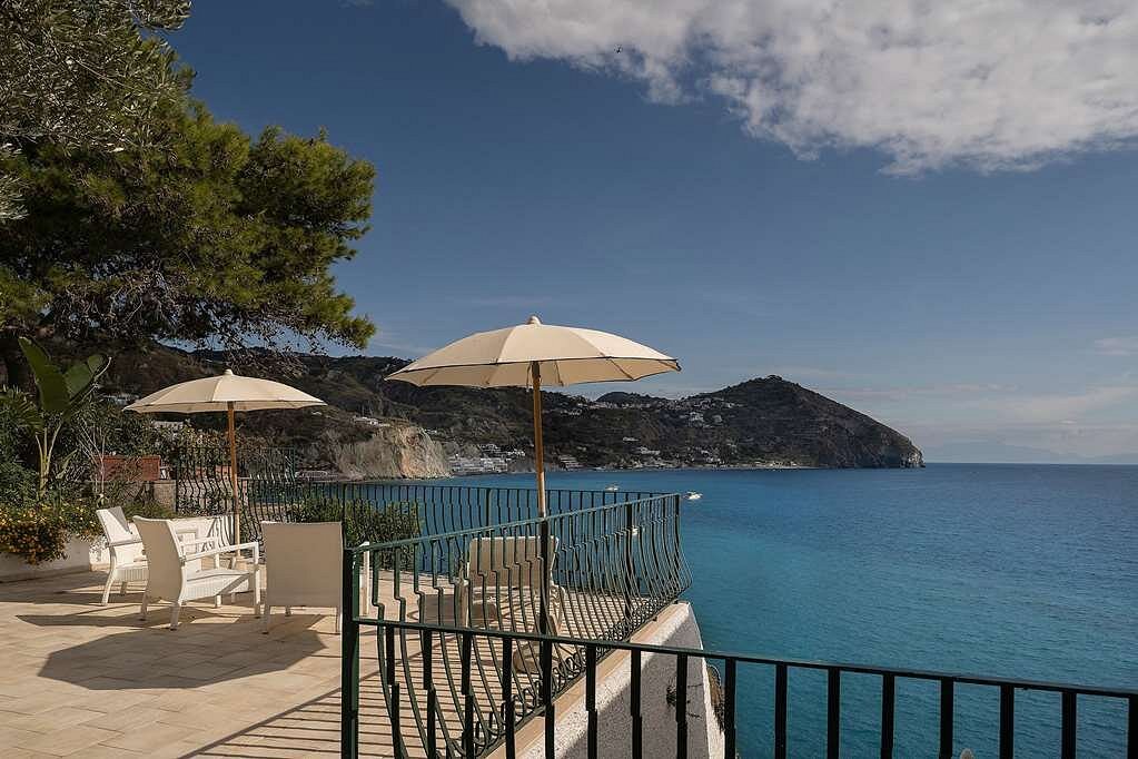 Seaside terrace with lounge chairs and umbrellas at Miramare Sea Resort in Ischia, Italy, overlooking the clear blue water and coastline.