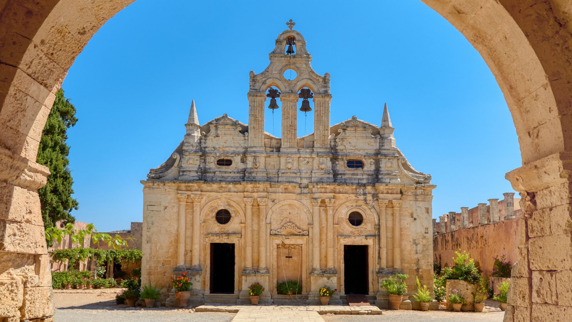 The facade of the Arkadi Monastery in Crete, featuring a stone structure with a bell tower and arched doorway, set against a clear blue sky.