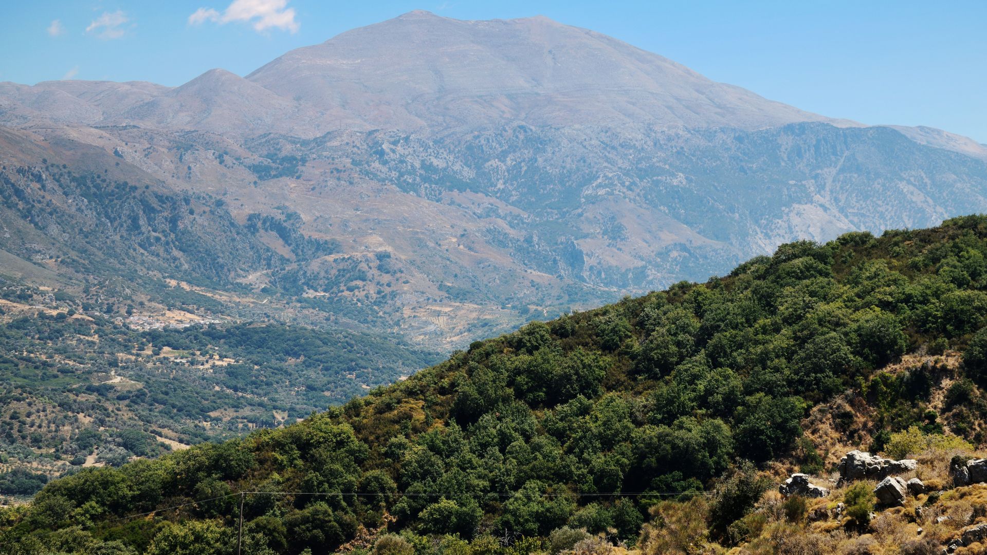 A landscape view of Mount Ida (Psiloritis) in Crete, with a foreground of green, tree-covered hills and the mountain rising in the background under a blue sky.