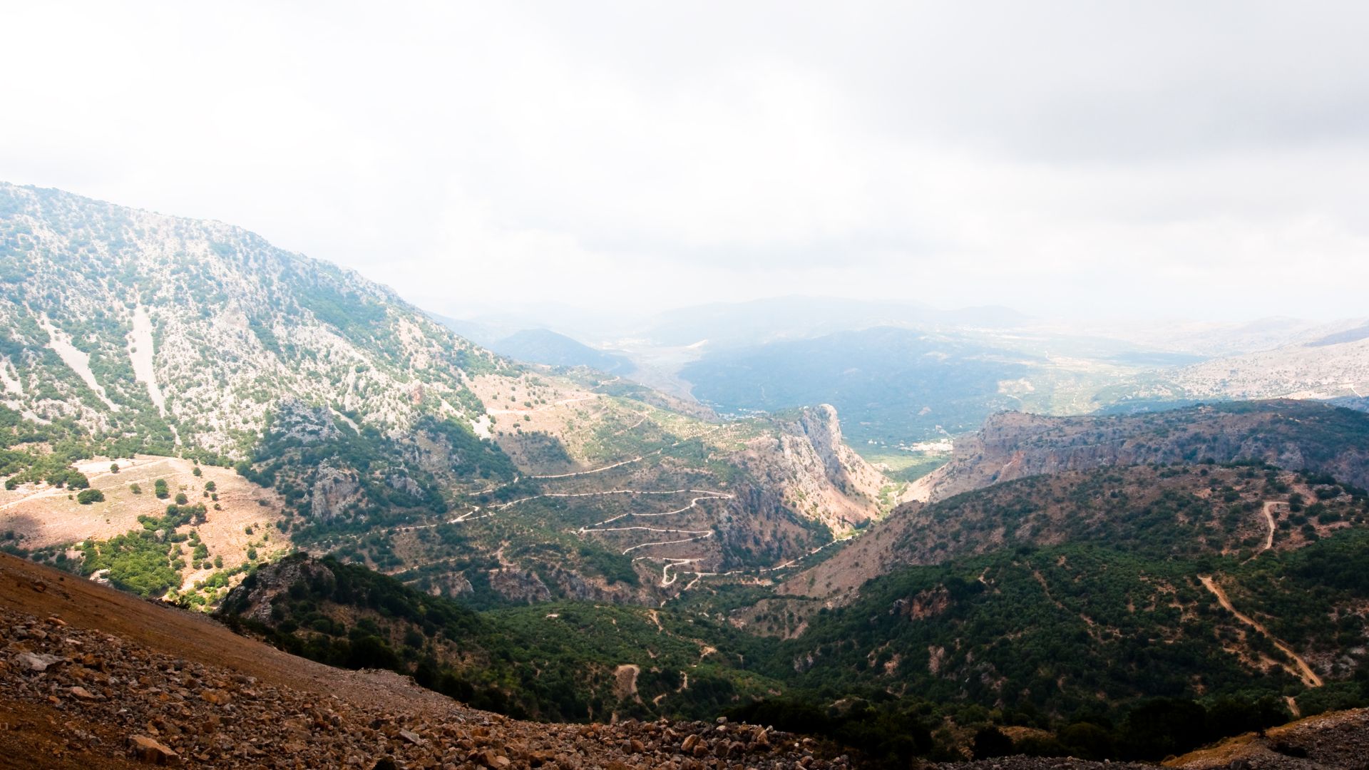 A scenic view of Mount Ida in Crete, featuring rugged terrain, winding roads, and lush greenery under a cloudy sky.