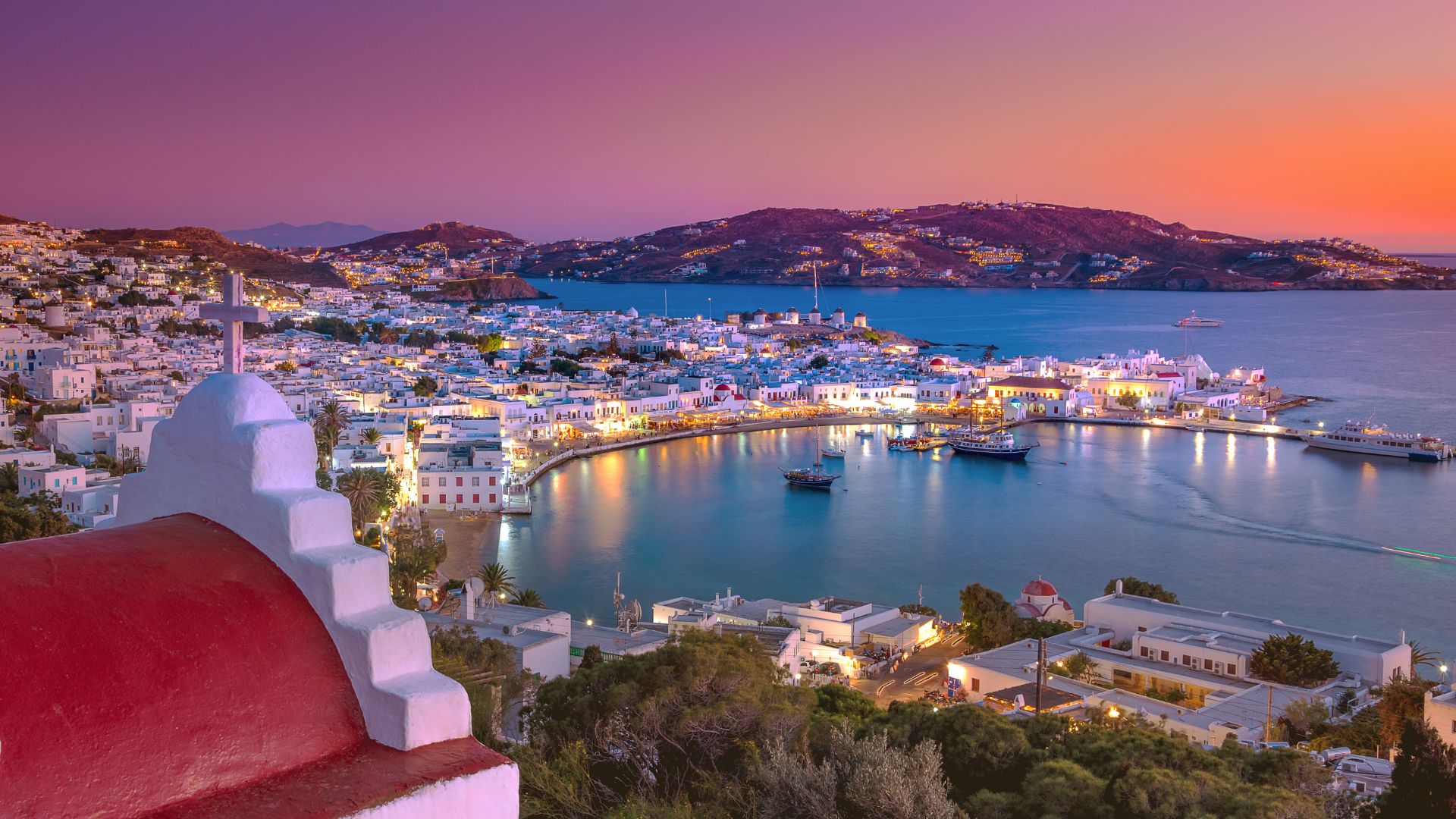 A scenic view of Mykonos town at dusk, showing white buildings, a harbor, and a colorful sky.