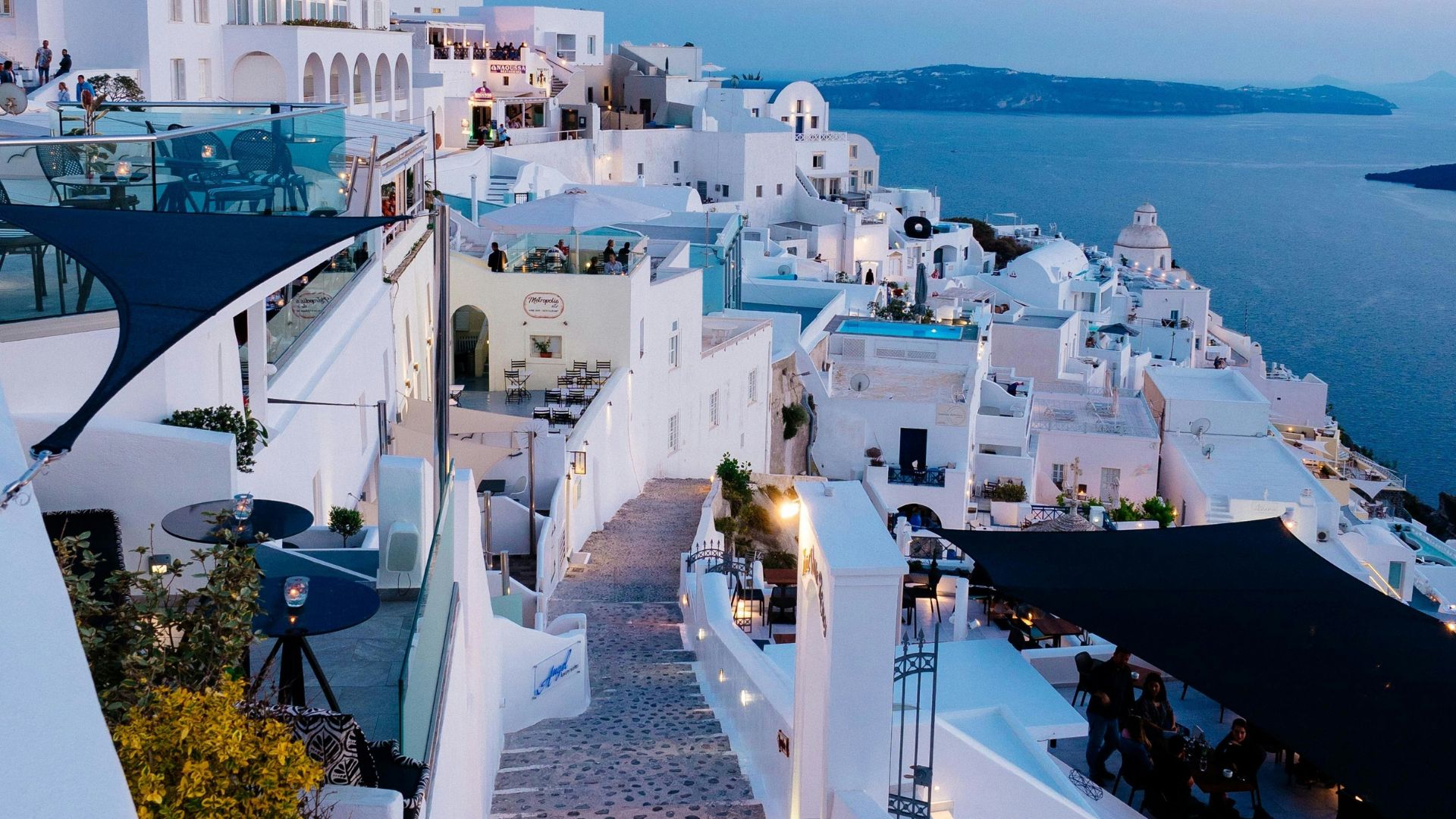 A narrow, cobblestone street in Mykonos Town with whitewashed buildings and a few trees.