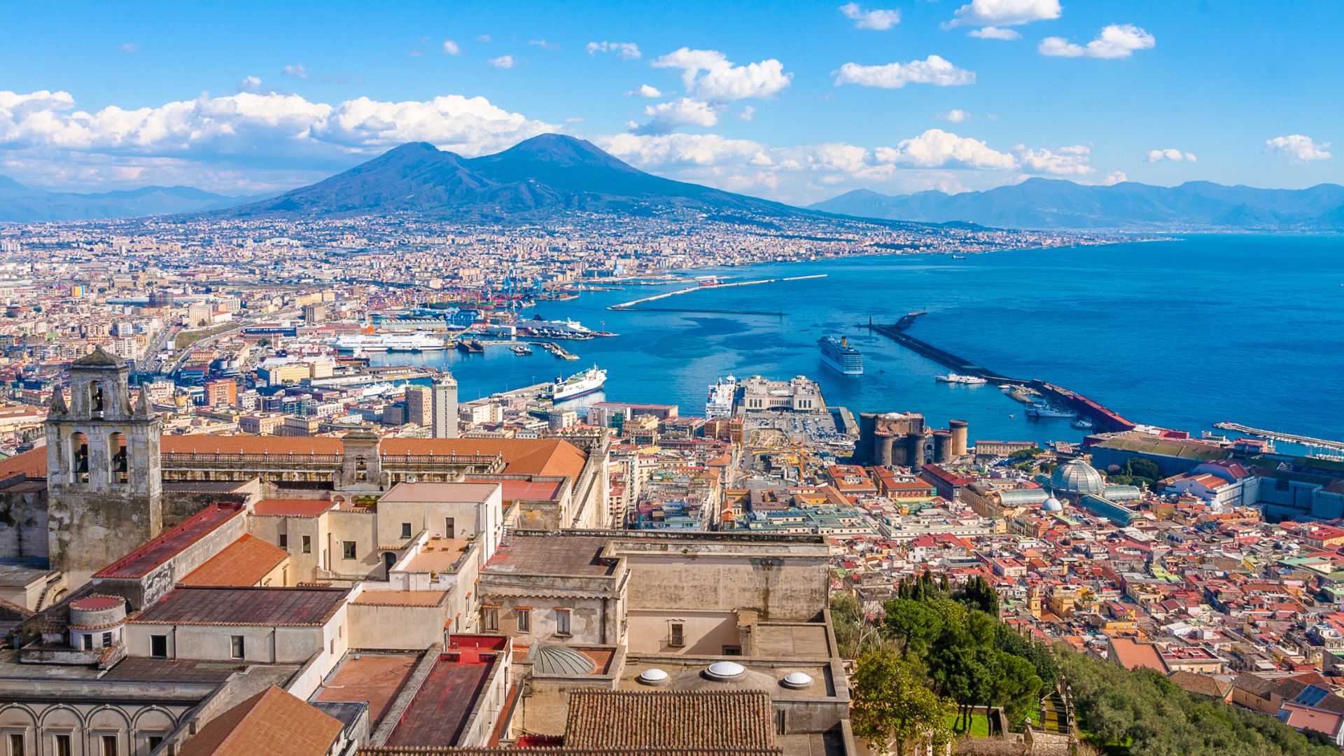 A panoramic view of Naples, Italy, showing the city sprawling along the coast of the Gulf of Naples with Mount Vesuvius in the distance.