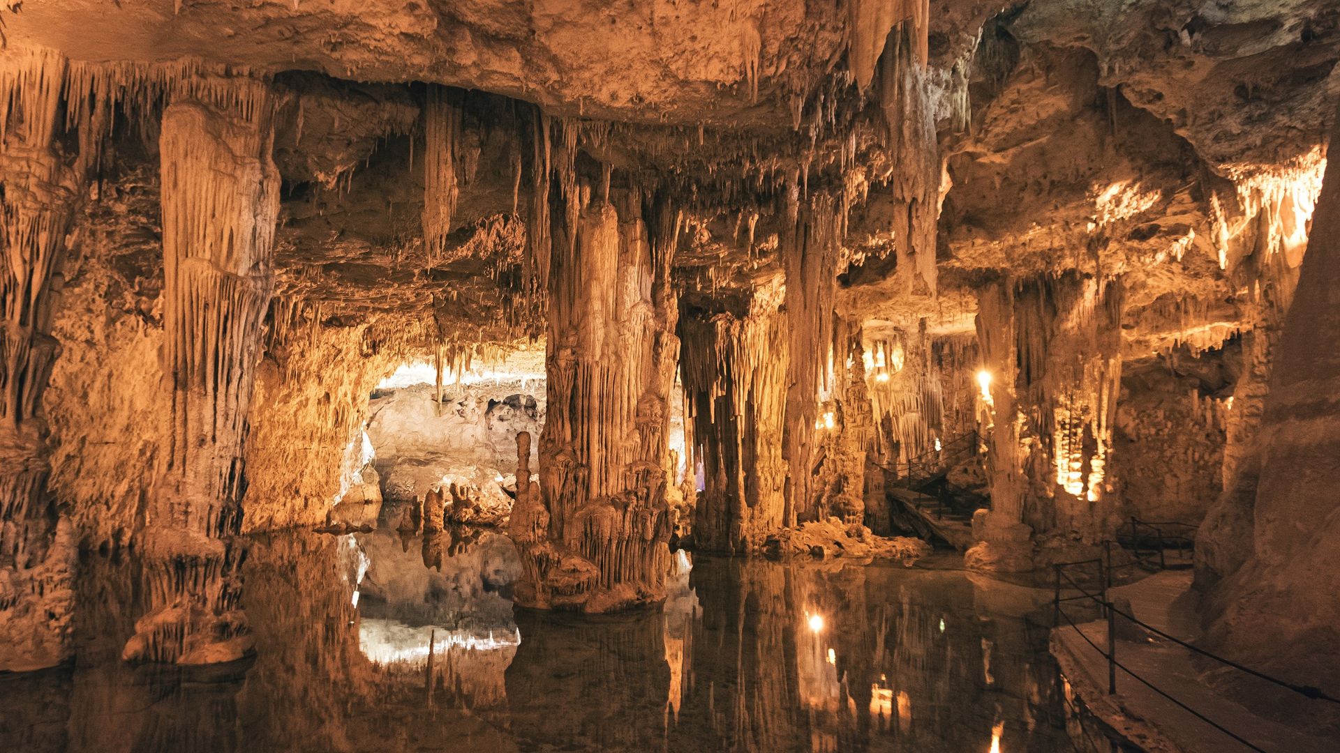 Inside Neptune's Grotto, stalactites and stalagmites are reflected in the clear waters of Lake La Marmora, illuminated by artificial lighting, with a walkway visible on the right.
