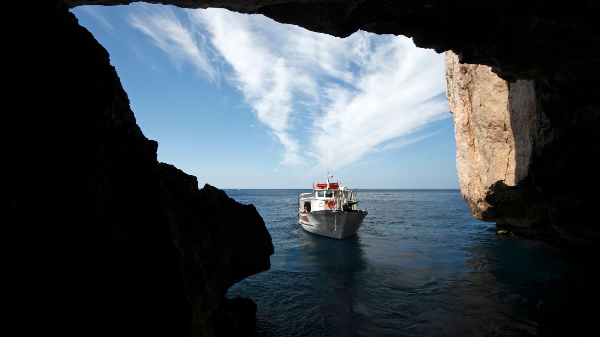 A boat approaches the entrance of Neptune's Grotto, a sea cave with a rocky opening, with a blue sky and wispy clouds visible above.