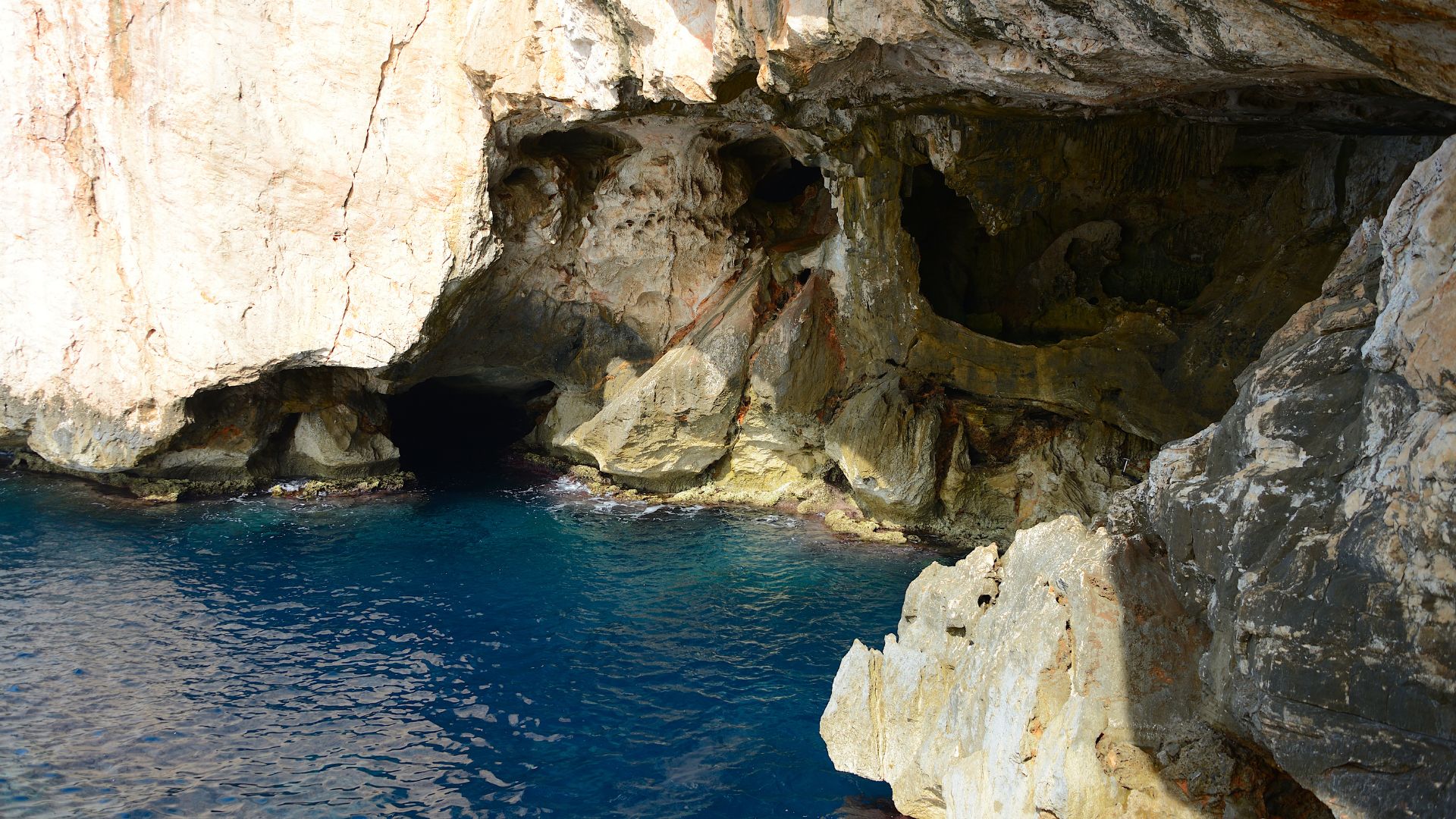 A wide, open space inside a cave with stalactites and stalagmites. The cave has a blue-ish color, likely from the sea that surrounds it. The cave appears to have an entrance and a staircase leading up.