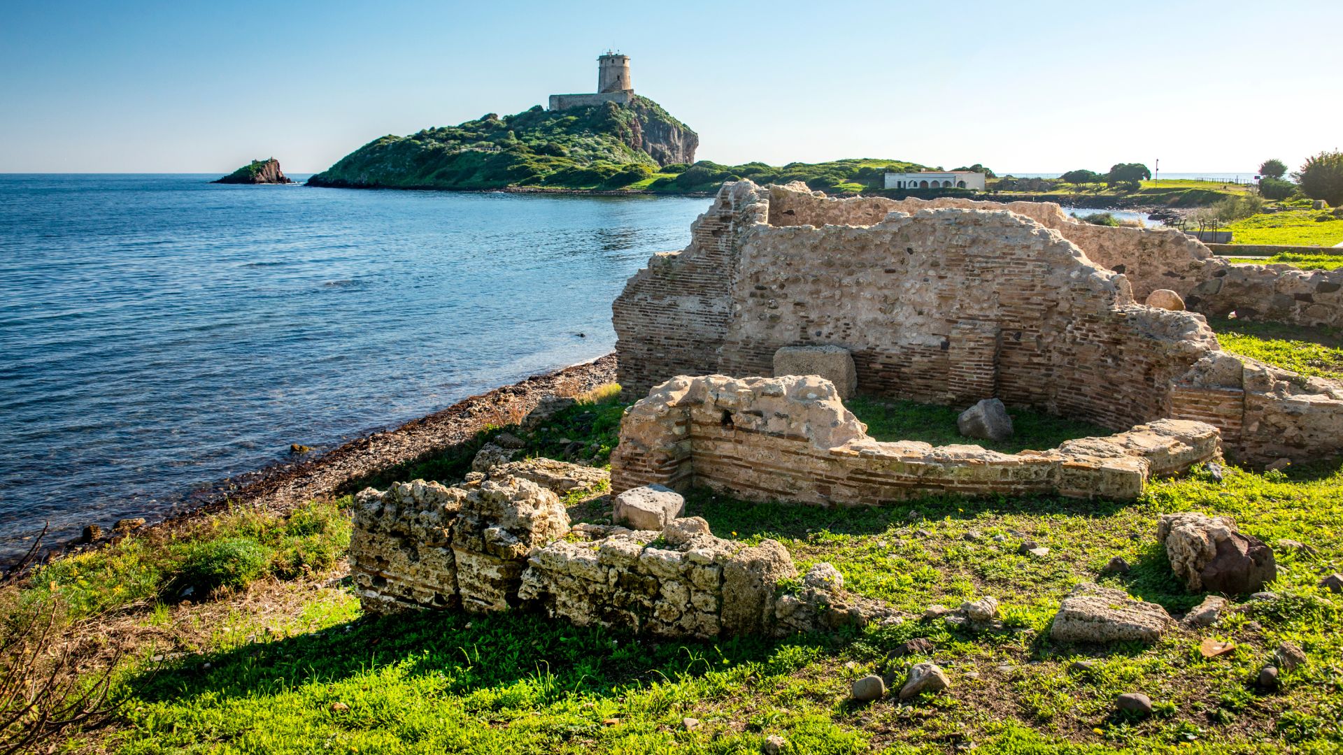 The ruins of Nora, an ancient city with stone structures, are set against the backdrop of the sea and a green hill with a tower on top.