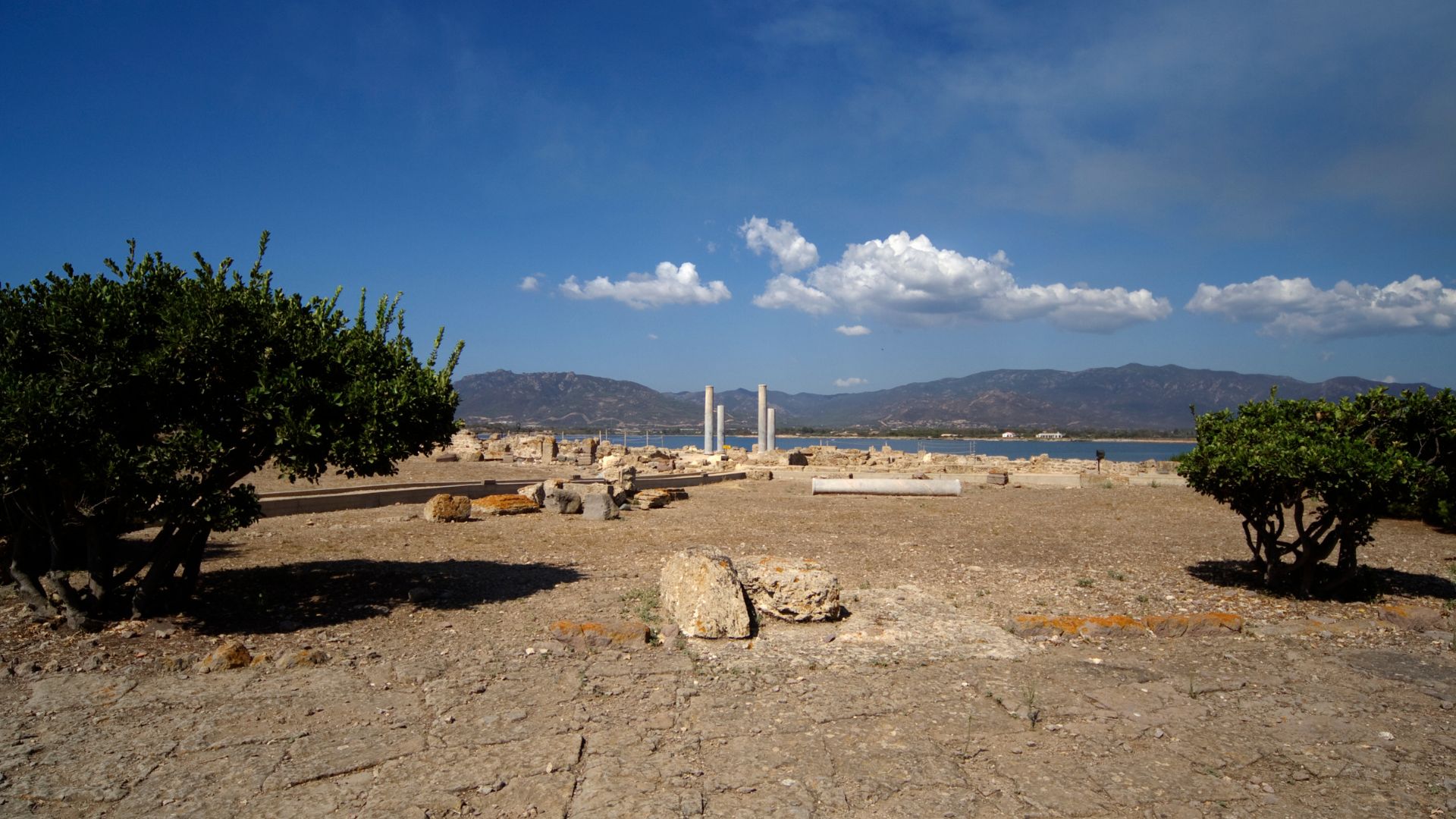 The ruins of the ancient city of Nora, with visible stone structures, columns, and the landscape of the Sardinian coast in the background, under a partly cloudy sky.