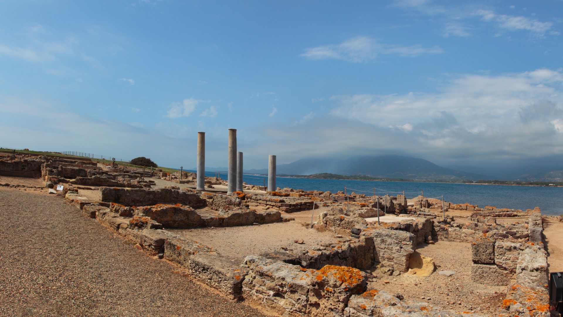 The ruins of the ancient city of Nora, with visible stone structures, columns, and the landscape of the Sardinian coast in the background, under a partly cloudy sky.