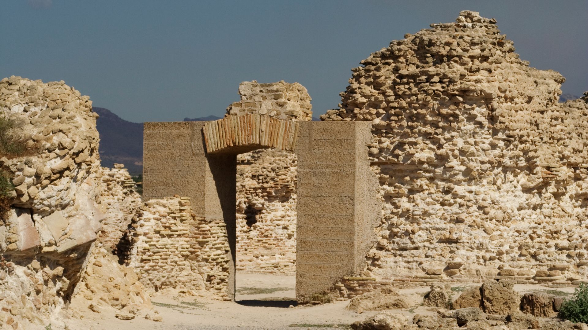 The ruins of the ancient city of Nora, featuring a stone archway and crumbling walls, set against a backdrop of a clear sky and distant hills.