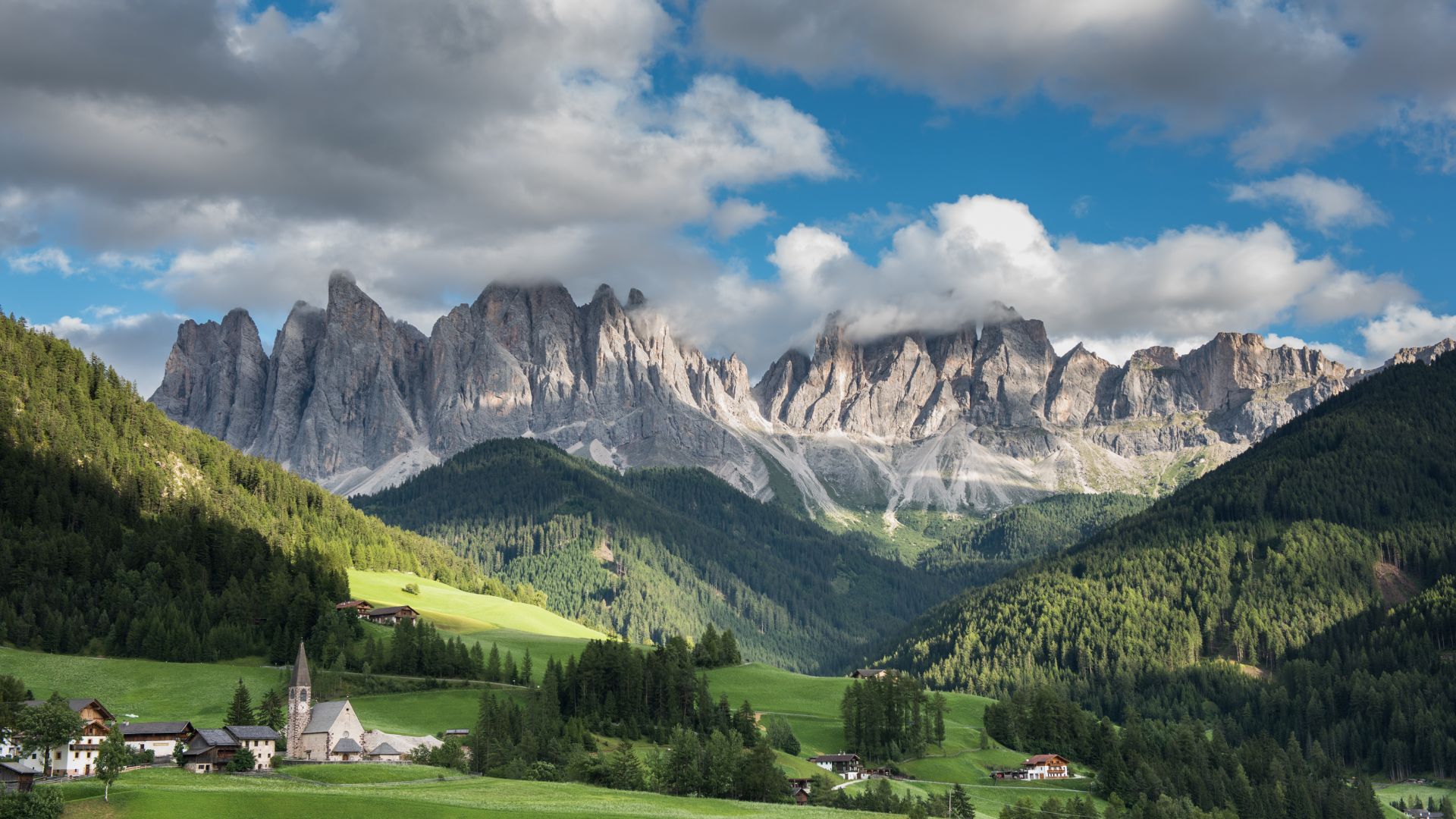 A scenic view of the Puez-Odle Nature Park in the Dolomites, featuring rugged mountain peaks, lush green valleys, and a small village nestled at the foot of the mountains under a partly cloudy sky.