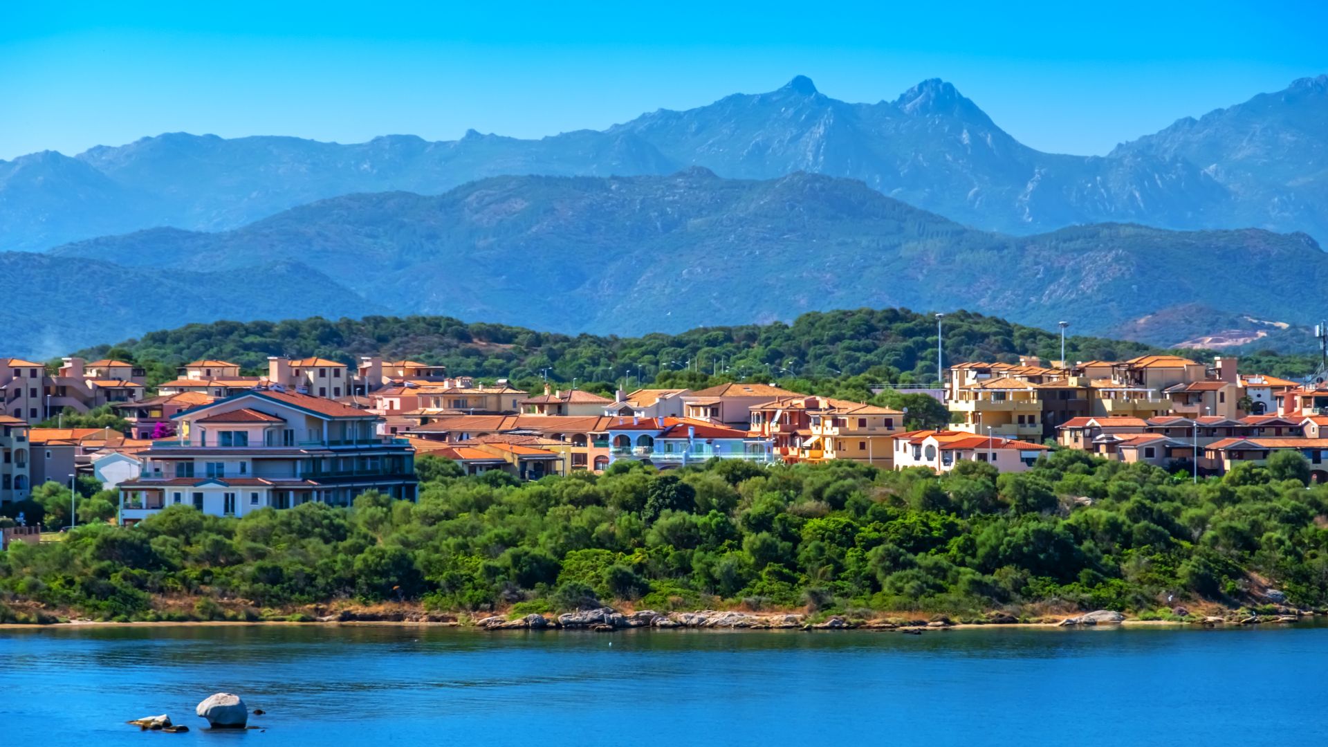 A scenic view of Olbia, Sardinia, with colorful buildings nestled among green trees and mountains in the background, bordering the blue Gulf of Olbia.