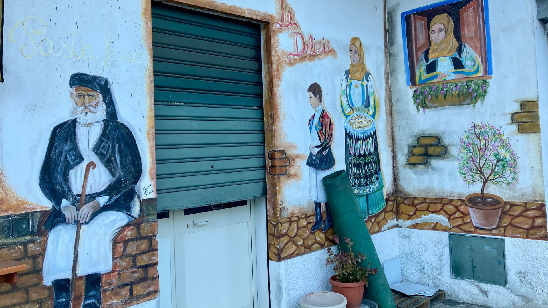 A mural depicting traditional Sardinian figures and scenes adorns the exterior of a building in Orgosolo, Sardinia. The artwork features an elderly man with a cane, a family in traditional dress, and a woman in a window, all painted on a textured wall alongside a closed shutter and potted plants.