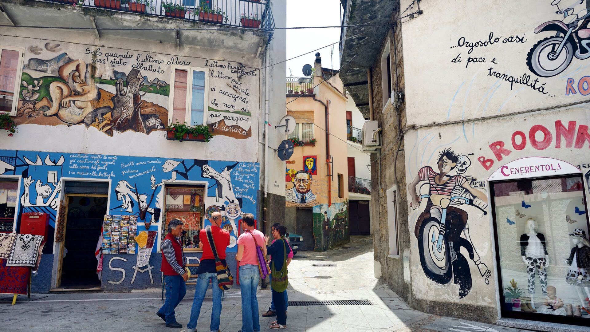 A street in Orgosolo, Sardinia, features murals on buildings, with a group of people standing in the foreground, observing the artwork. The murals depict various scenes, including social and political commentary, reflecting the town's history of protest and resistance. 