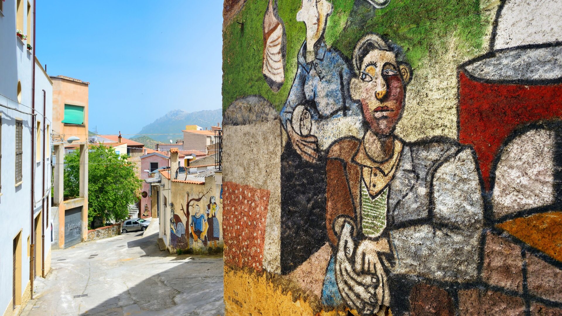 A close-up of a mural in Orgosolo, Sardinia, Italy, depicting several figures in a rough, folk art style, with a street scene visible in the background. The mural is characterized by its bold lines and earthy colors, showing people in what appears to be a social or daily life setting. 