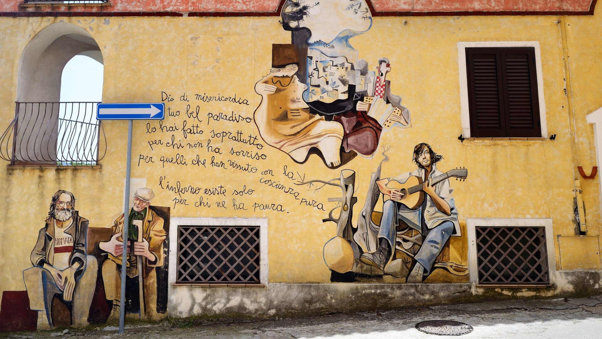A colorful mural adorns the exterior wall of a building in Orgosolo, Sardinia, Italy. The mural depicts several scenes and figures, including seated men, a guitarist, and abstract representations of people and landscapes. Text is incorporated into the mural in Italian, featuring phrases such as "Dio di misericordia il tuo bel paradiso lo hai fatto soprattutt per chi non ha sorriso per quelli che han vissuto con la coscienza pura" and "l'inferno esiste solo per chi ne ha paura," which translate to "God of mercy, your beautiful paradise you made especially for those who have not smiled, for those who have lived with a pure conscience" and "hell exists only for those who are afraid." The mural is painted on a yellow wall with architectural elements such as windows, a balcony, and a doorway visible. 