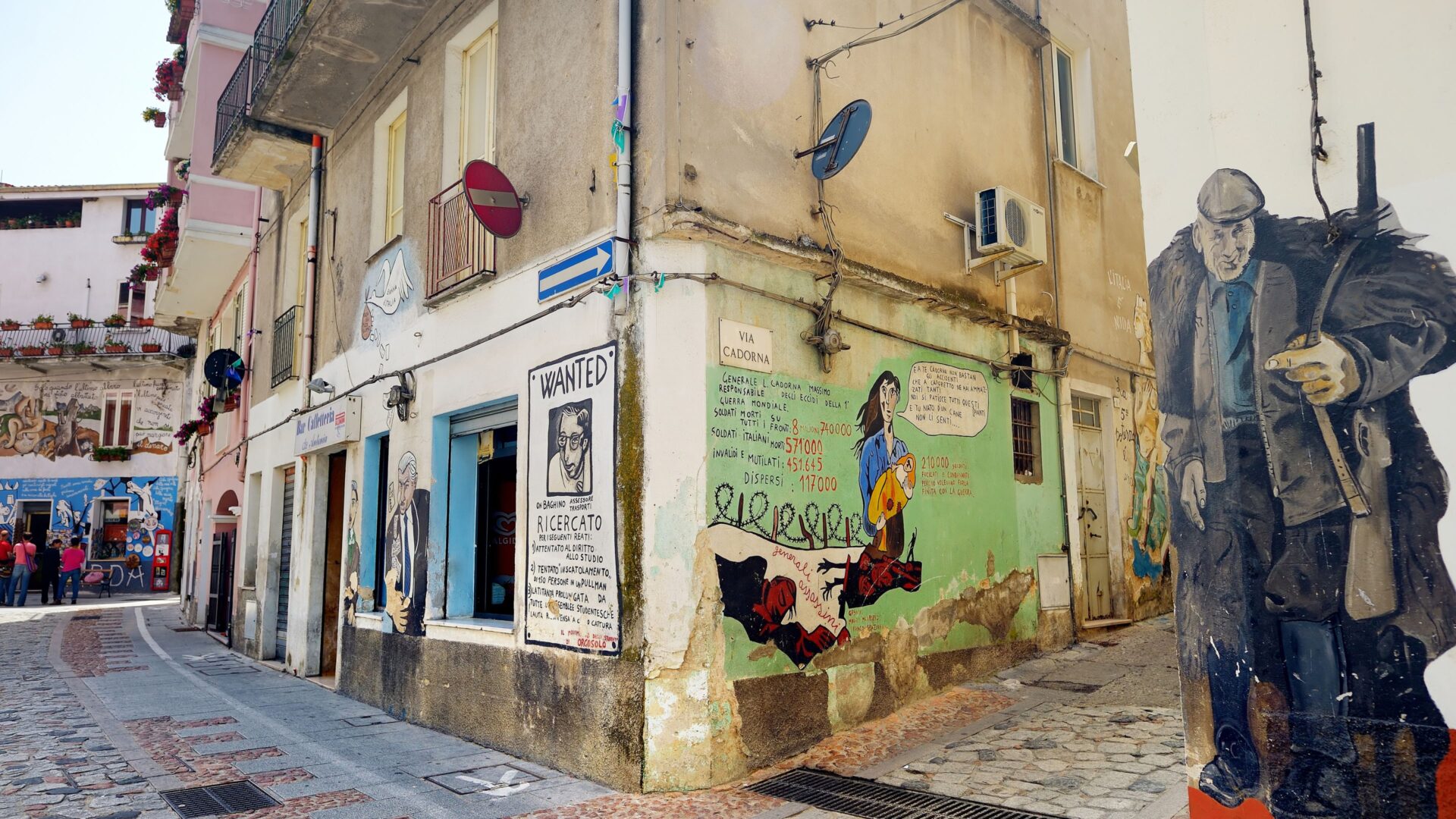 A street corner in Orgosolo, Sardinia, features murals on the walls of buildings, including a "Wanted" poster and a painting depicting figures and text. The artwork reflects the socio-political themes often found in the town's murals, with a focus on local and global issues, and the text on the wall includes what appears to be a phone number.