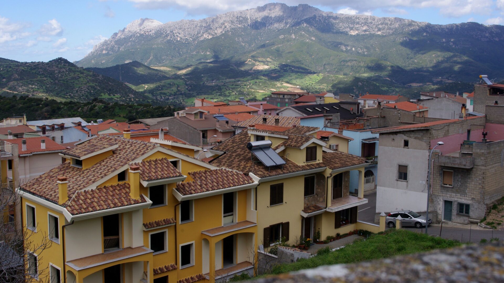 A view of a town with terracotta-roofed buildings nestled in a valley, with a large mountain in the background. The buildings are primarily yellow and orange, contrasting with the green hillsides and the gray, rocky peak of the mountain. 