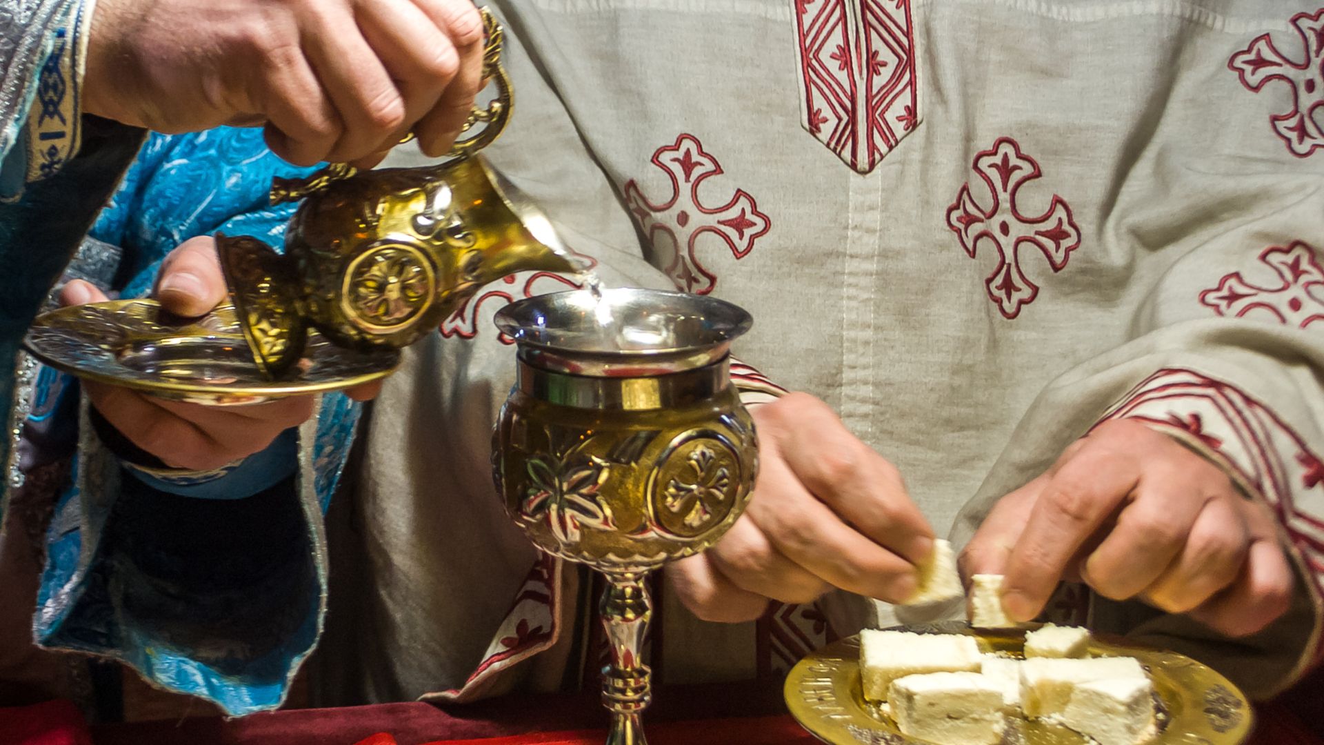 The image shows a priest performing the Eucharist during an Orthodox Liturgy. The priest is pouring wine from a golden vessel into a chalice, while preparing pieces of bread on a plate.