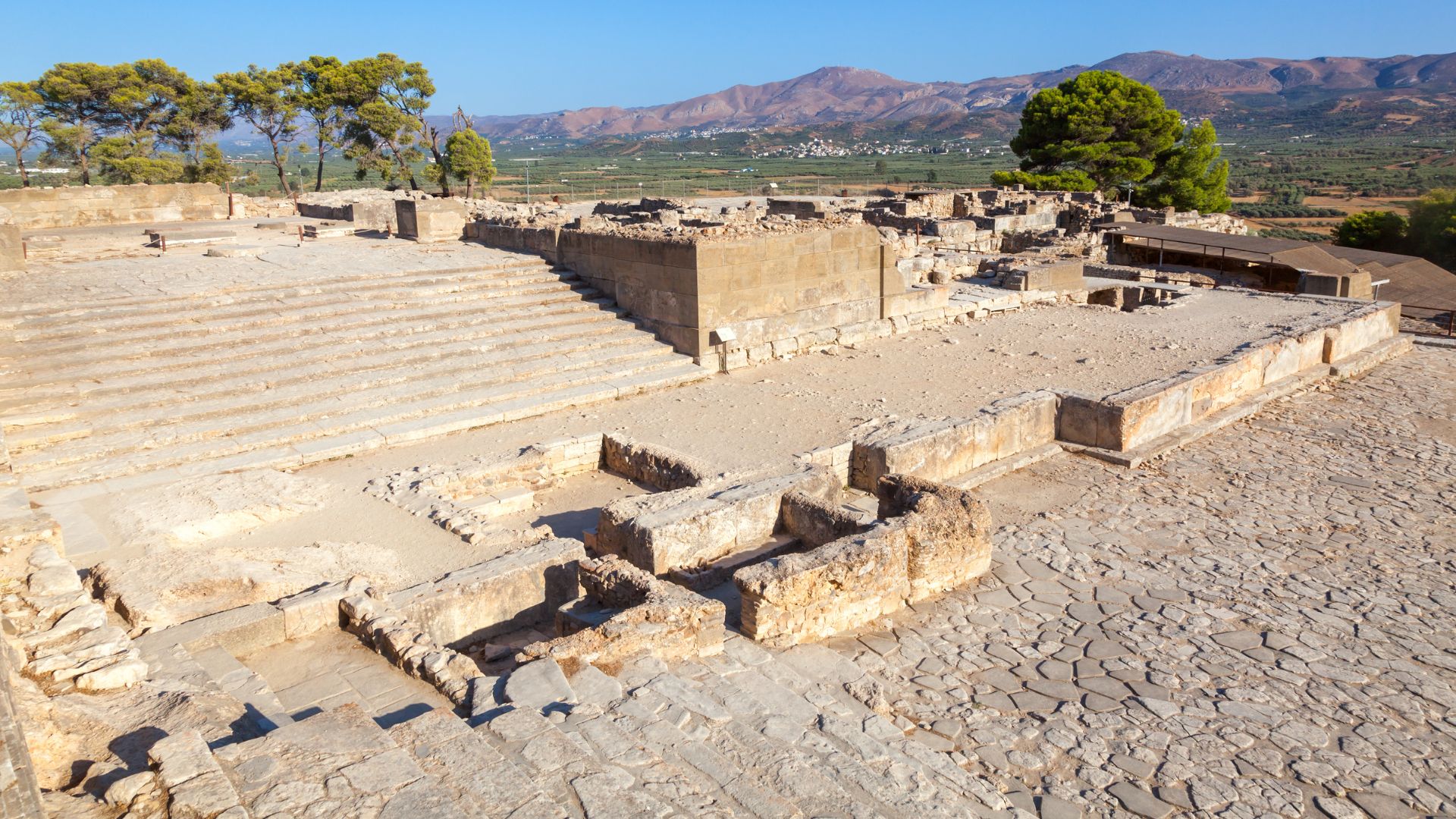 Aerial view of the ruins of the Palace of Phaistos, an ancient Minoan site in Crete, featuring stone walls, steps, and the surrounding landscape.