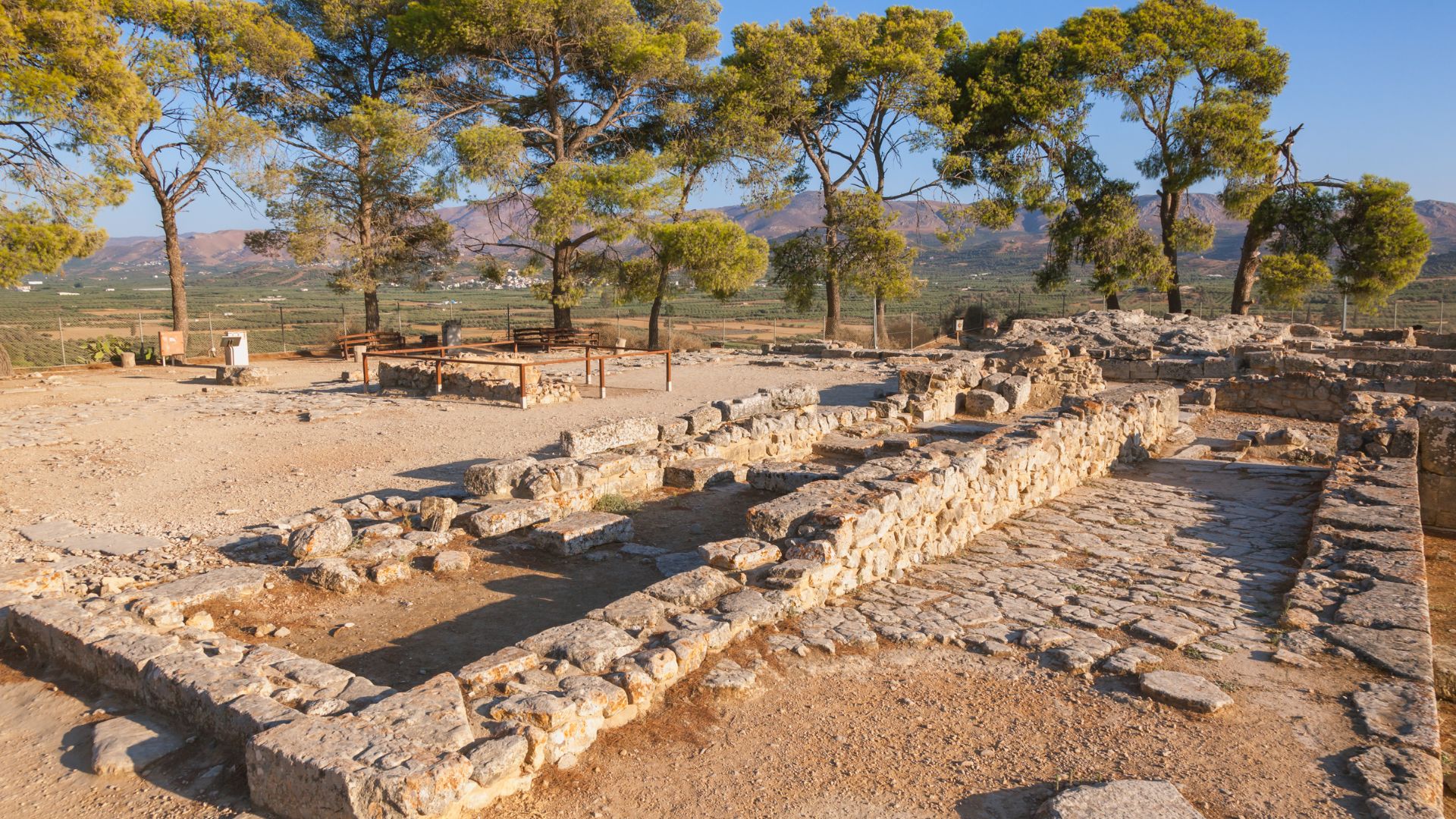 Ruins of the ancient Palace of Phaistos, with stone walls and paved areas, set against a backdrop of trees and distant mountains in Crete, Greece.