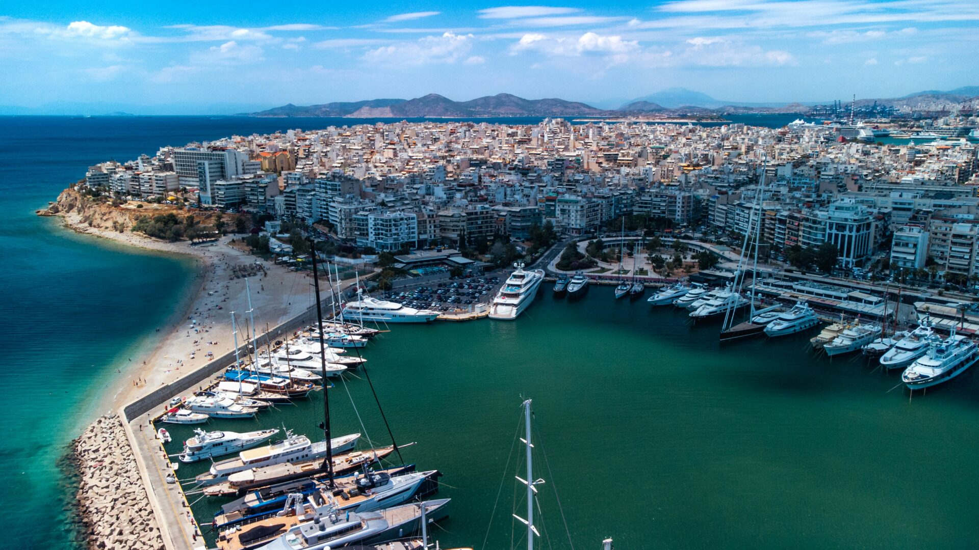 Aerial view of Piraeus Harbour in Greece with yachts docked and a beachside cityscape in the background.