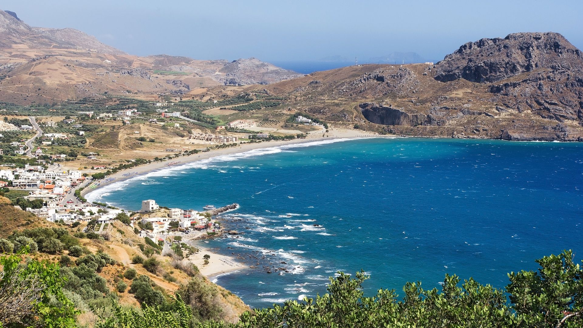 Scenic view of Plakias, Crete, Greece, featuring a crescent-shaped sandy beach, clear blue water, and mountainous terrain.