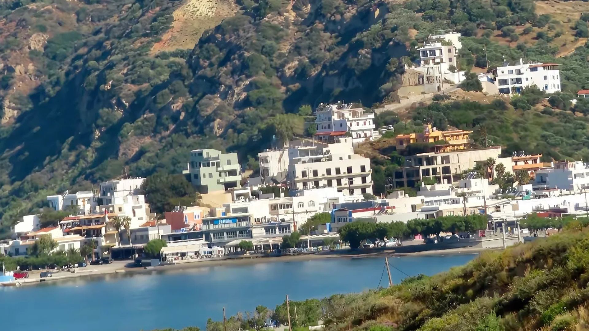 Plakias in Crete, Greece nestled on a hillside with white buildings, lush greenery, and a blue bay.