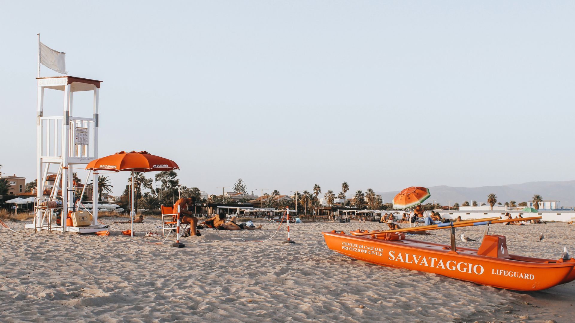 A sunny day at Poetto Beach, featuring a lifeguard tower, orange umbrellas, people relaxing on the sand, and a bright orange lifeguard boat in the foreground, with the coastline and distant hills visible in the background.